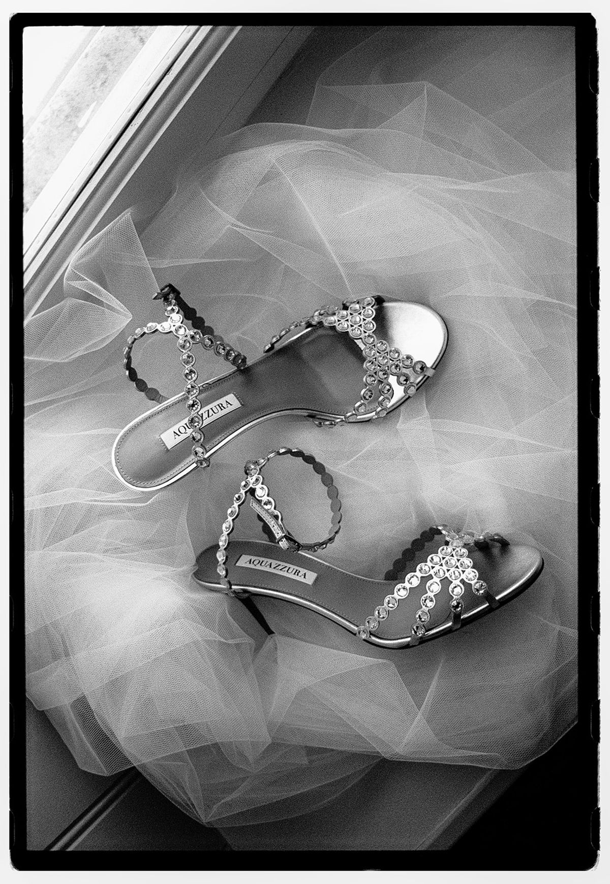 A pair of jewelled strappy sandals lies on soft tulle in black and white, framed by a dark film border.