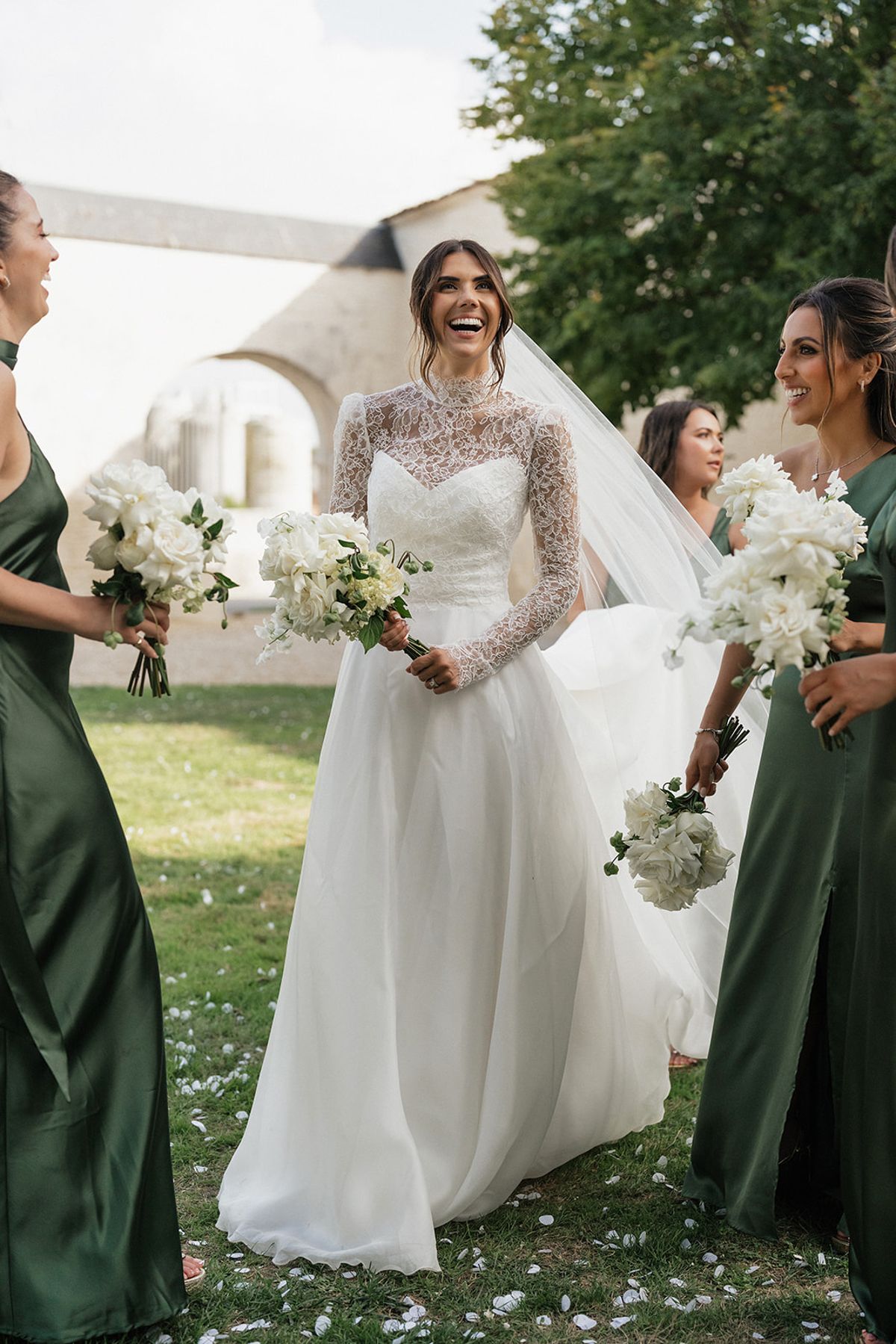A bride in a lace gown laughs while bridesmaids in green dresses holding white bouquets stand around her on the grass.