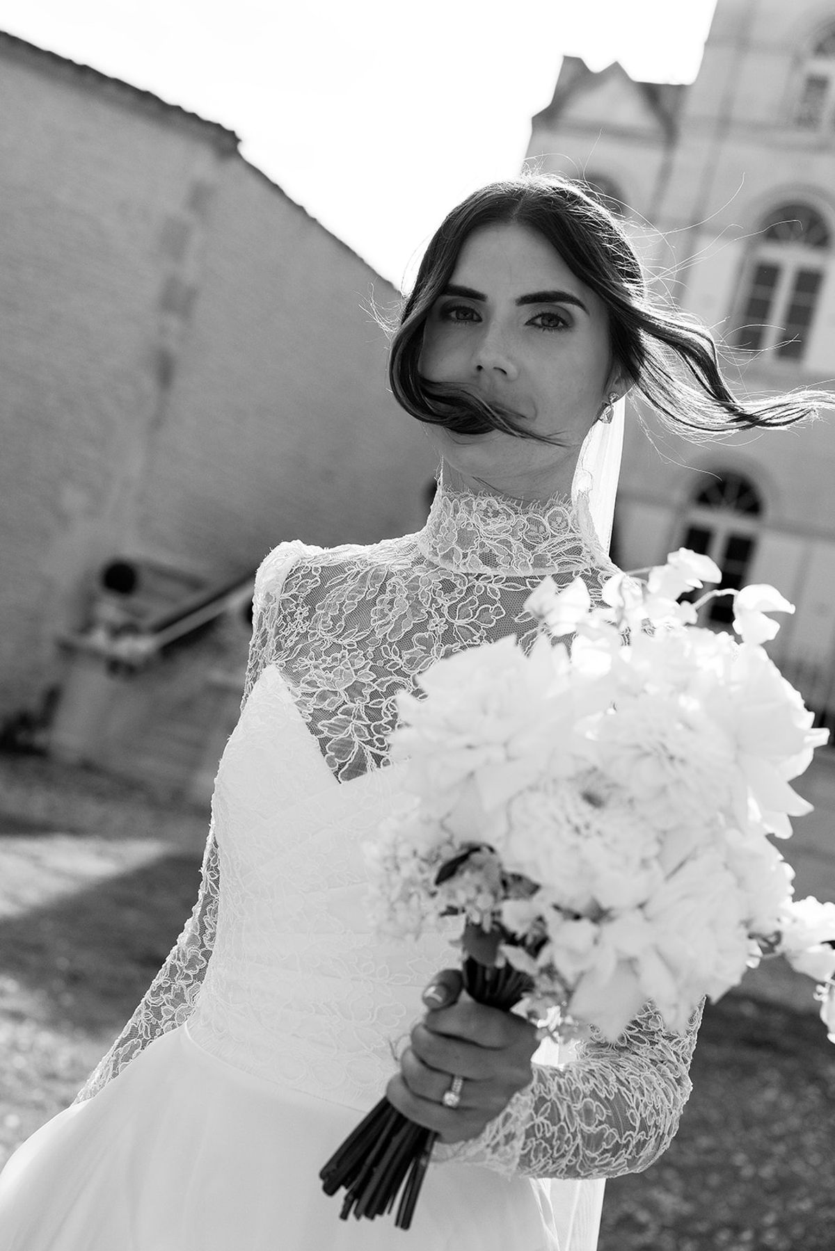 A black and white portrait of a bride in a high-necked lace gown holding a large white bouquet, her veil blowing across her face.