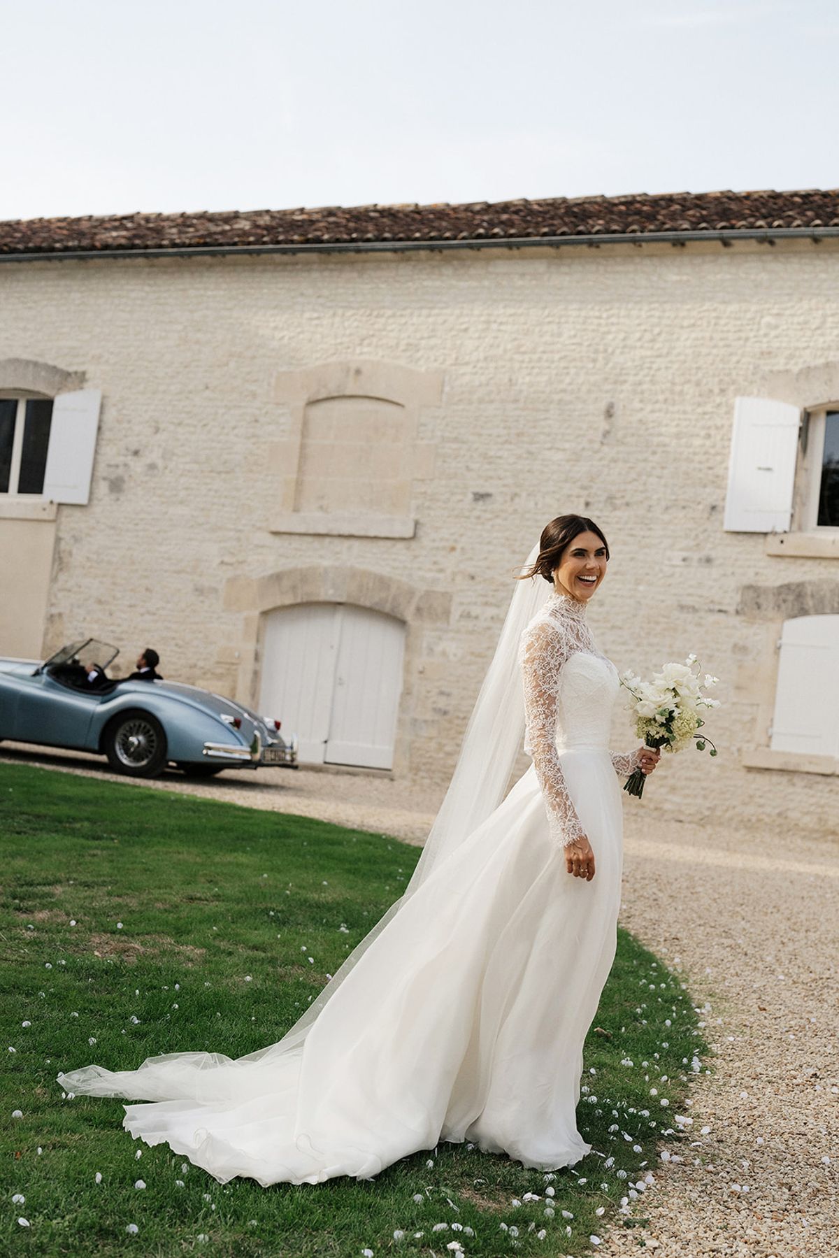 The bride in a lace-sleeved gown with a long veil walks across a lawn, holding a bouquet, with a vintage blue car behind.
