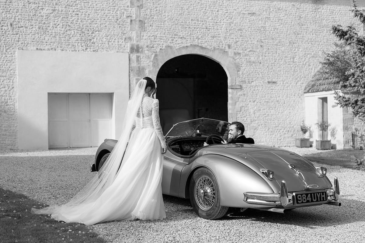 In black and white, the bride in a long veil stands beside a vintage open-top car where the groom sits, before a stone building.