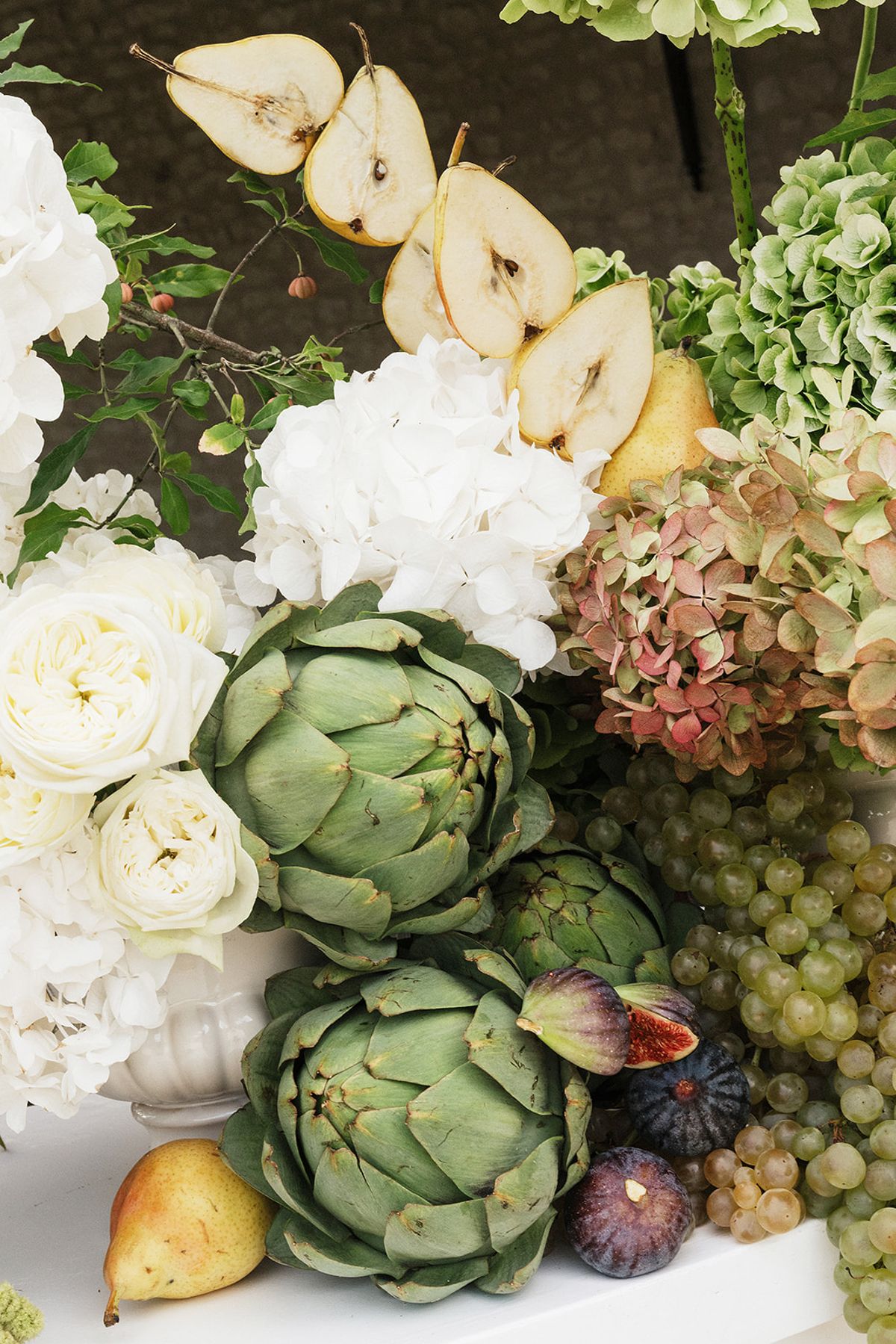 A centrepiece of white roses and hydrangeas is arranged with artichokes, sliced pears, grapes and figs.