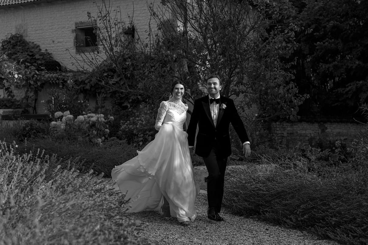 In black and white, the couple walk side by side along a garden path with lavender and a stone house behind.
