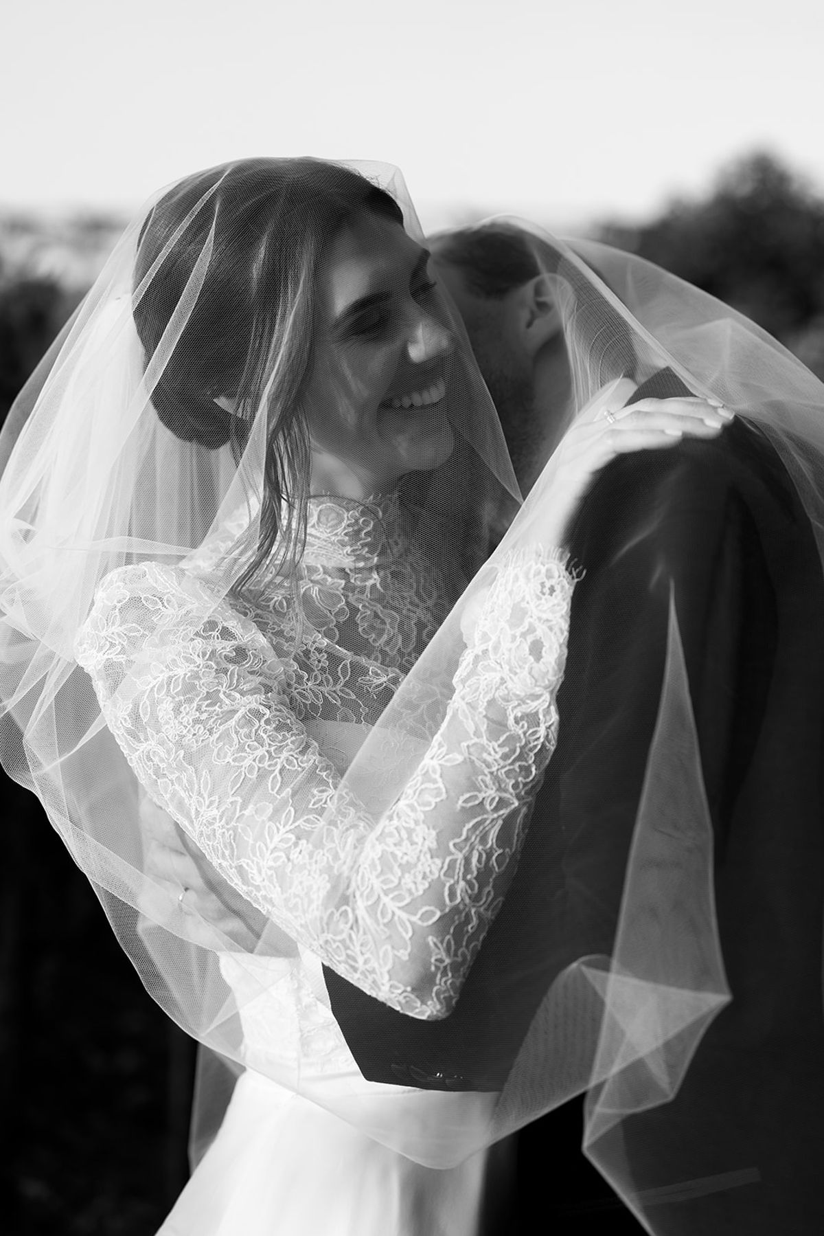 The bride, smiling, embraces the groom closely with her veil draped over them both, in black and white.