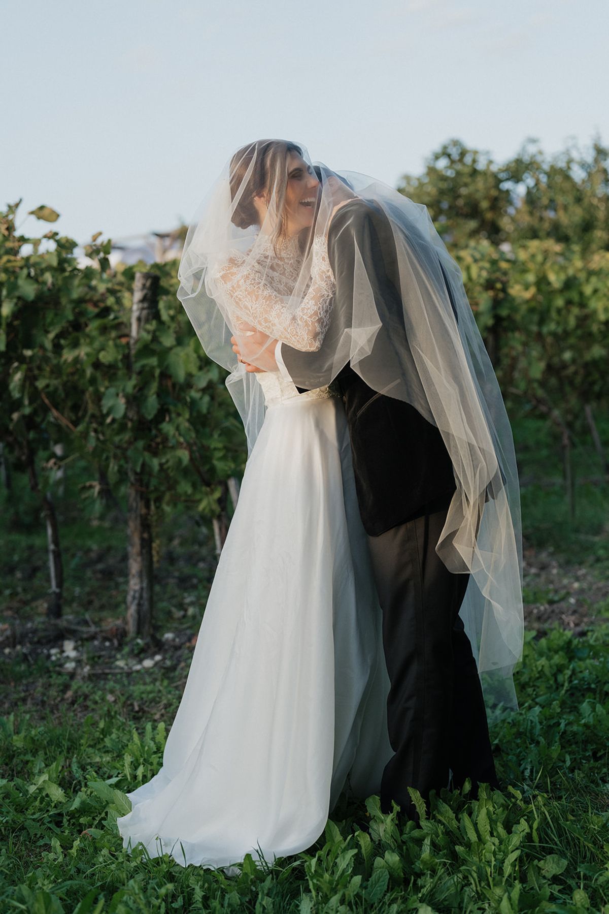 The couple embrace and kiss beneath the bride's lace veil in a vineyard, with rows of vines behind them.