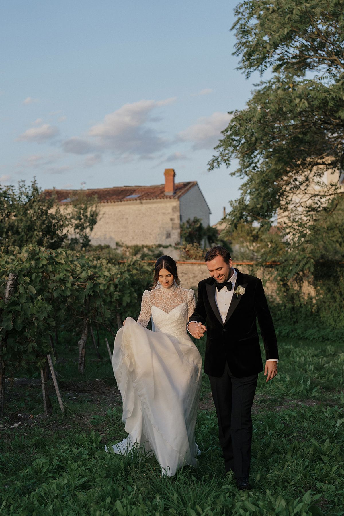 The couple walk arm in arm between rows of vines, the bride lifting her skirt, with a stone house and trees behind.