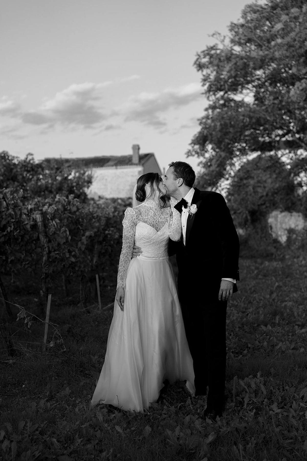 The bride in a lace-sleeved gown and the groom in a dark suit stand close together among vines, in black and white.
