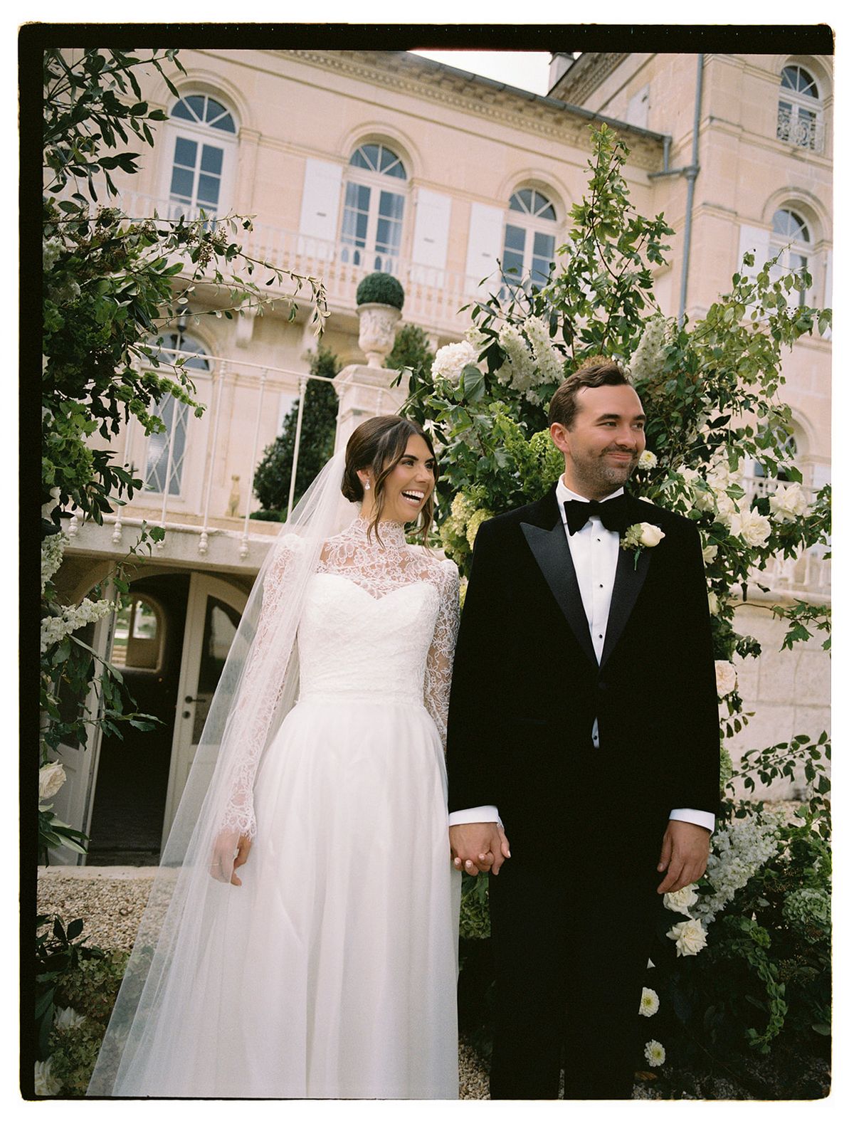 The couple stand holding hands in front of a pink-toned chateau, the bride in a lace-sleeved gown with a long veil.