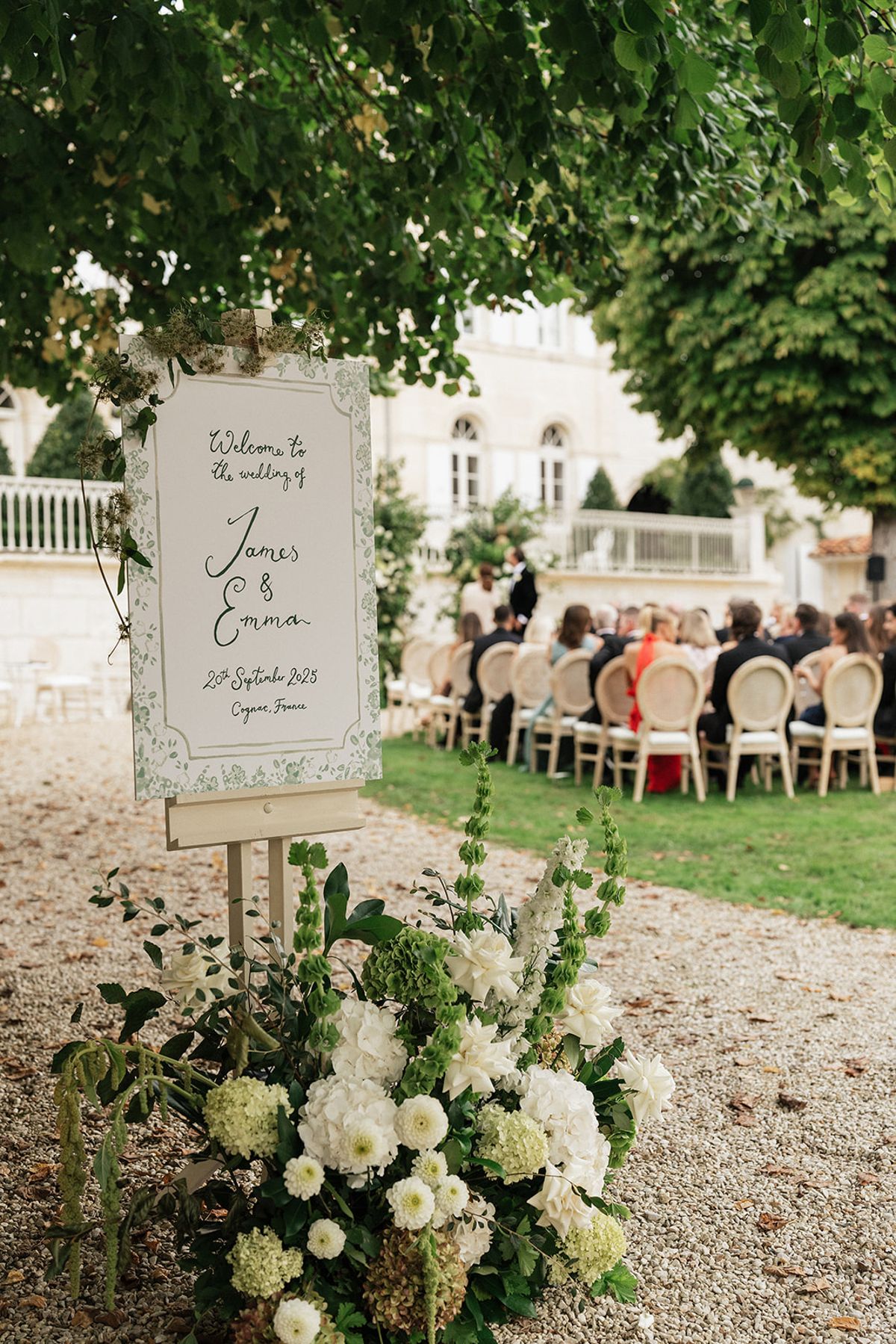 A framed welcome sign on an easel stands among white flowers, with rows of seated guests on a lawn in the background.
