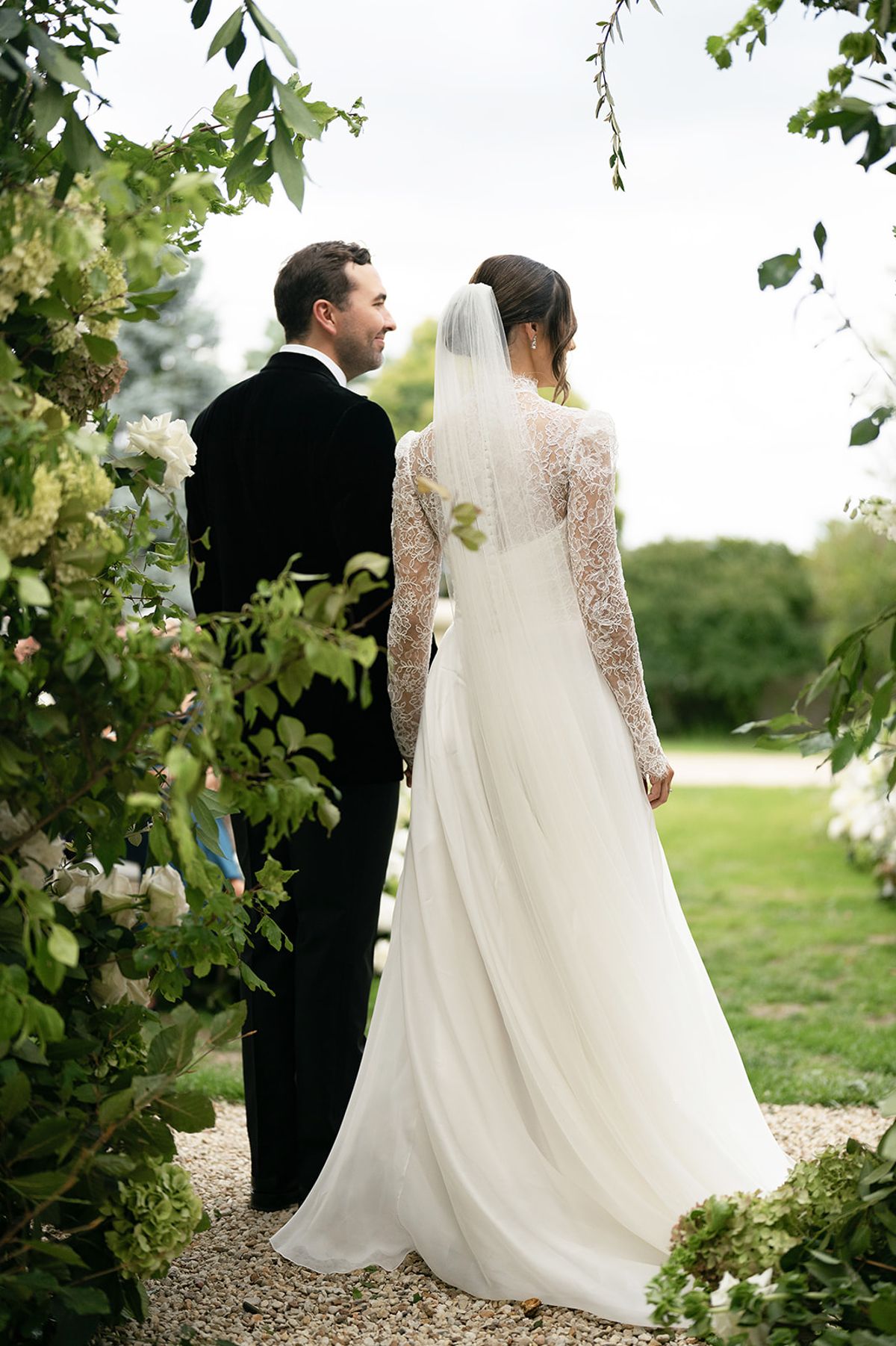 The bride in a long veil and lace-sleeved gown stands beside the groom in a dark suit, both facing a leafy garden.