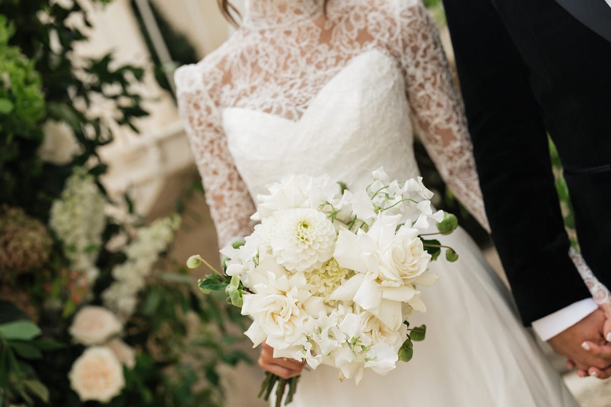 A close view of a bride's white bouquet and lace bodice, with the groom's dark sleeve and their joined hands beside her.