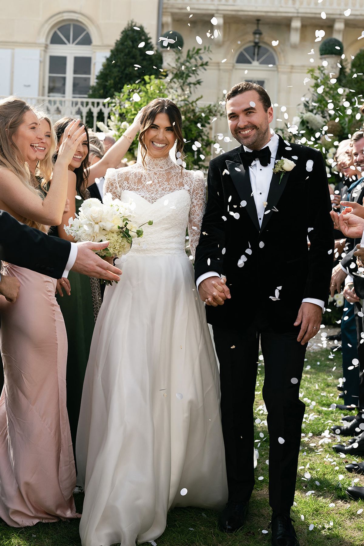 The couple walk hand in hand through a shower of petals while guests in green and pink dresses cheer around them.