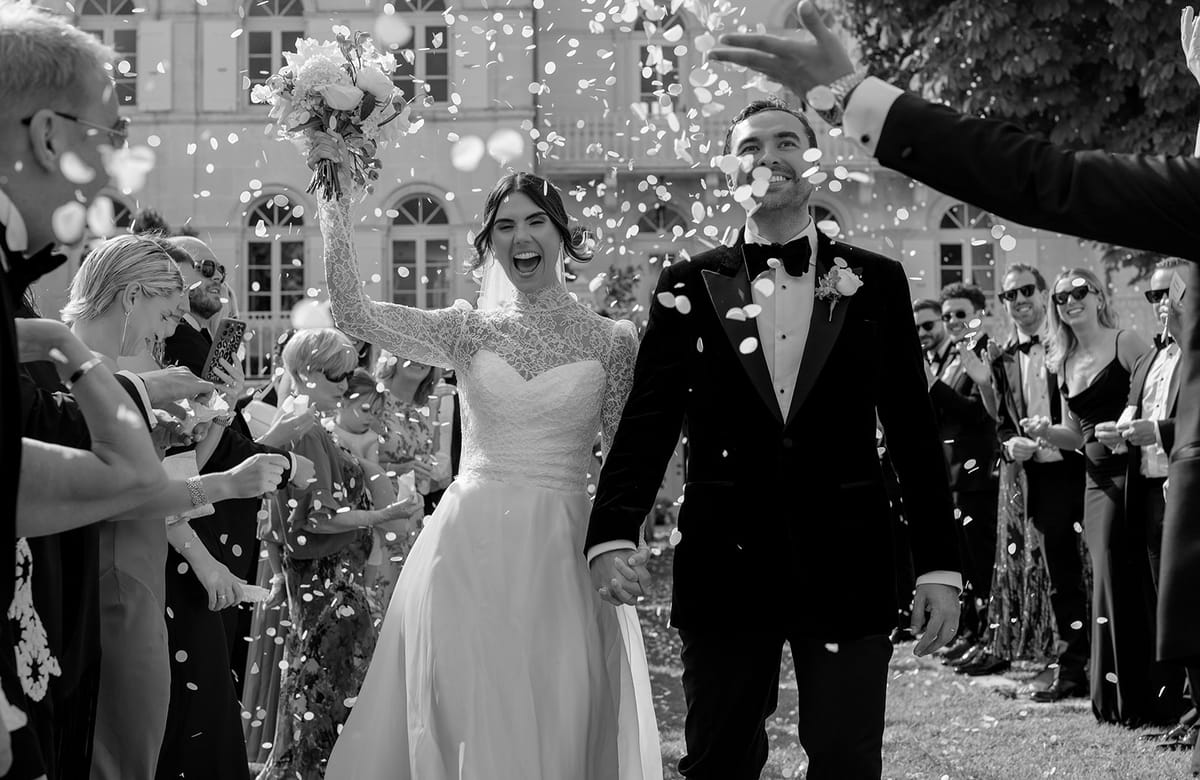 In black and white, the smiling couple walk between lines of guests as confetti falls, with a grand building behind.