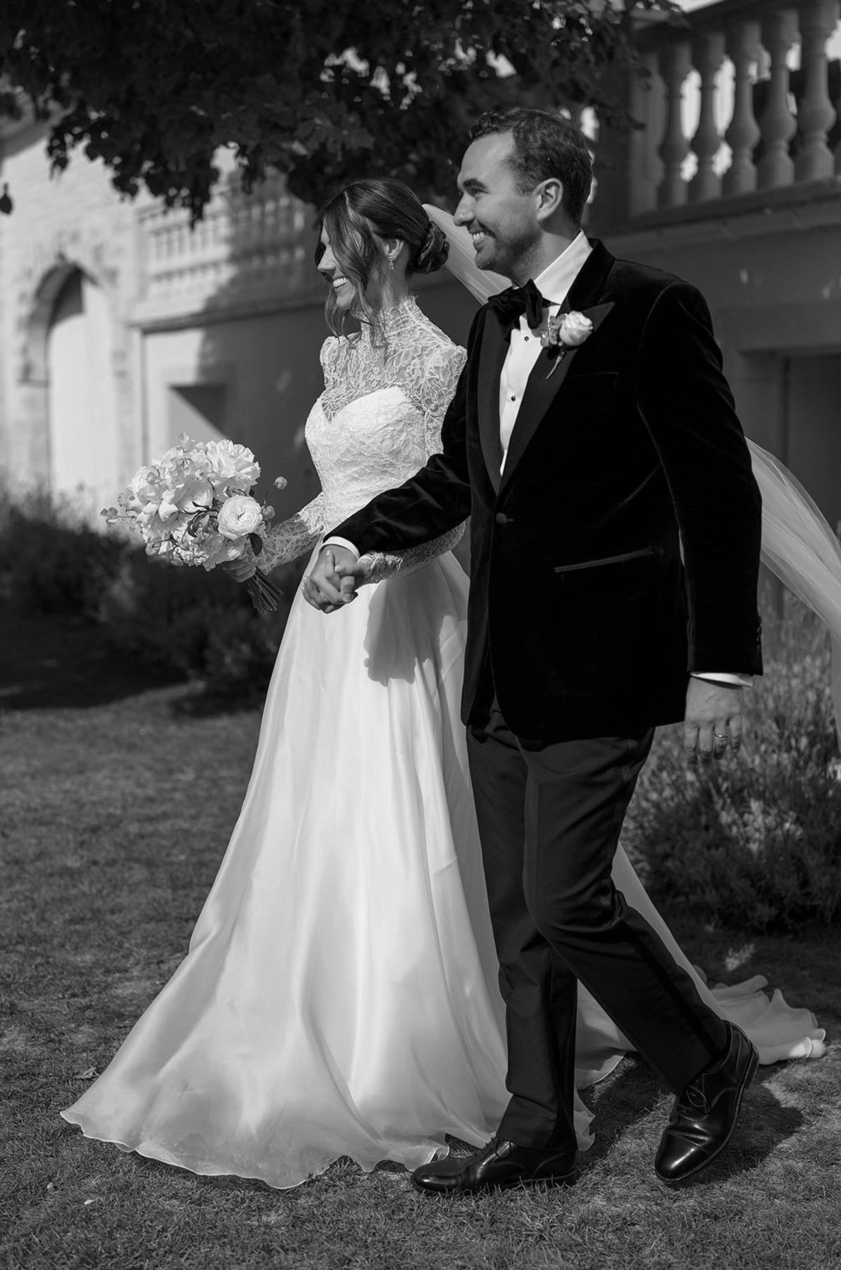 In black and white, the groom in a velvet jacket and the bride in a lace gown walk hand in hand, smiling.