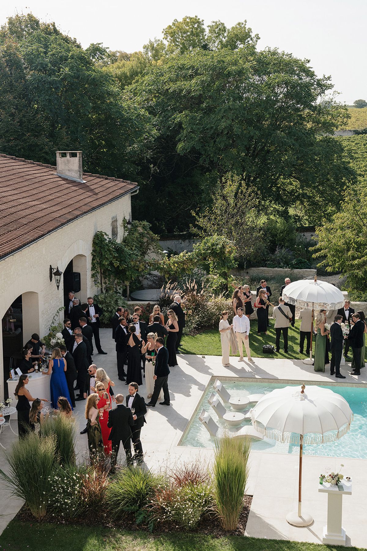 From a high viewpoint, dressed-up guests gather on a terrace beside a pool with white fringed parasols and a tiled-roof building.