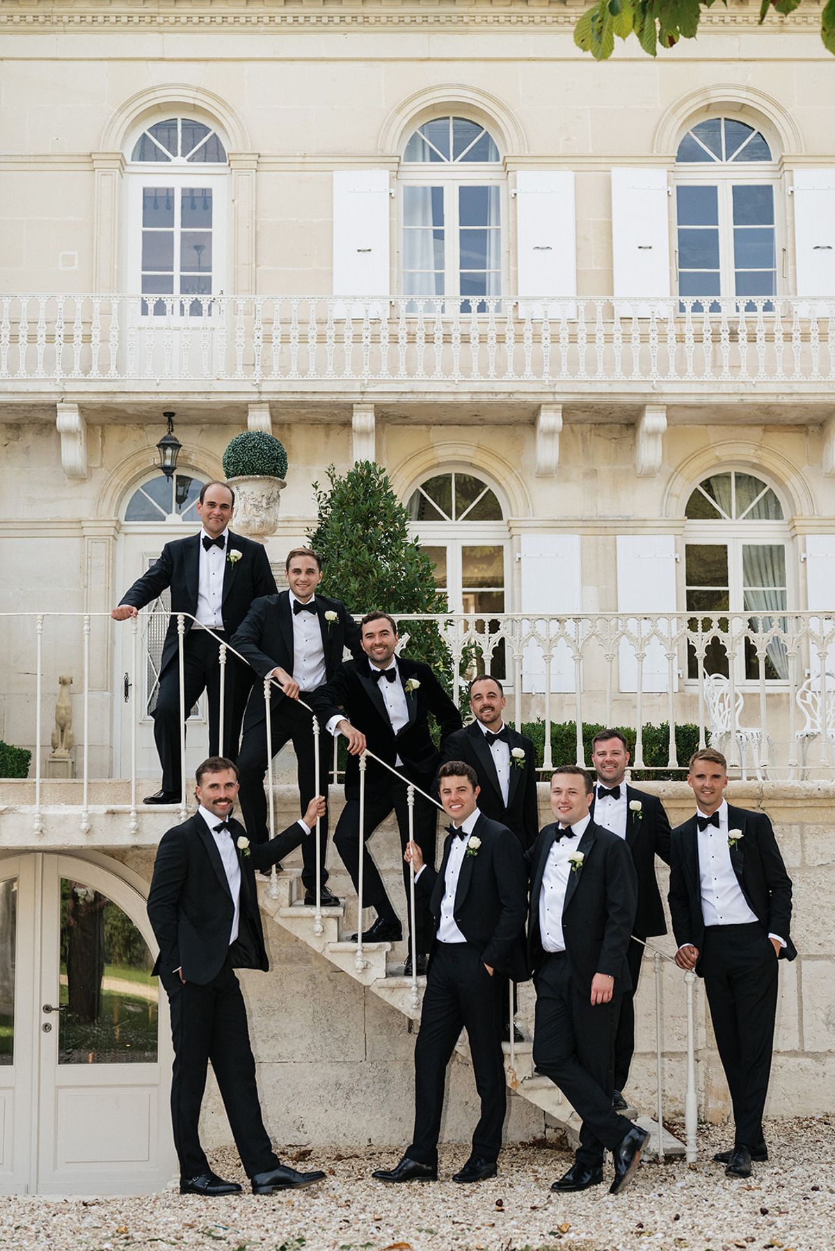 A group of men in black tuxedos pose on a white stone staircase in front of a chateau.
