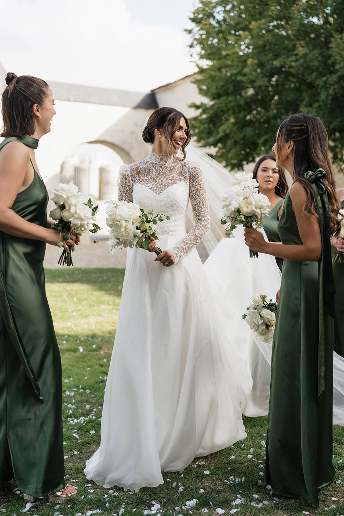 A bride in a lace gown walks across the grass between two bridesmaids in green satin dresses, all holding white bouquets.