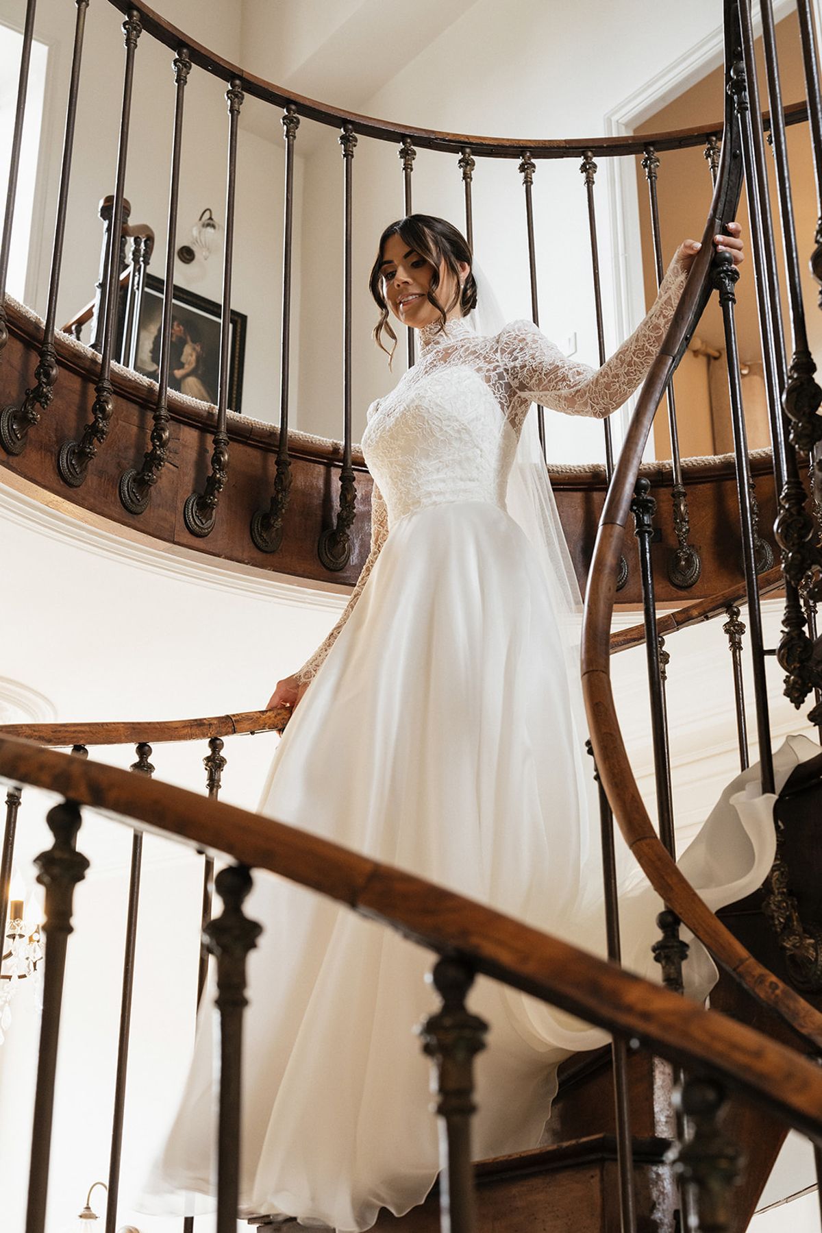 A bride in a long-sleeved lace gown with a full skirt walks down a spiral staircase with a dark wrought-iron banister.