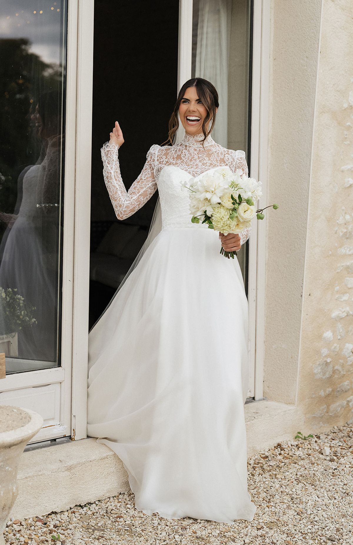 A smiling bride in a high-necked lace gown holds a white bouquet as she stands in an open glazed doorway beside a stone wall.