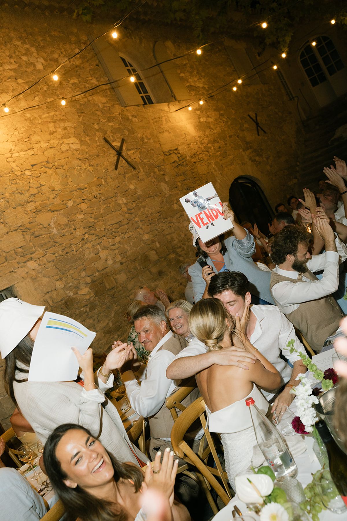 The bride and groom embrace as guests cheer at the tables after dark