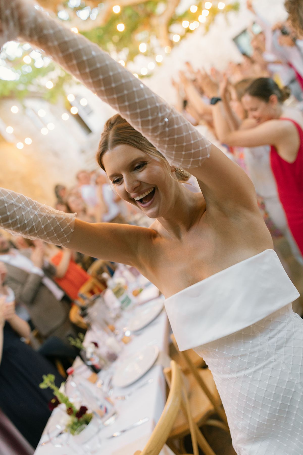 The bride raises her arms and laughs as guests celebrate at the dinner tables