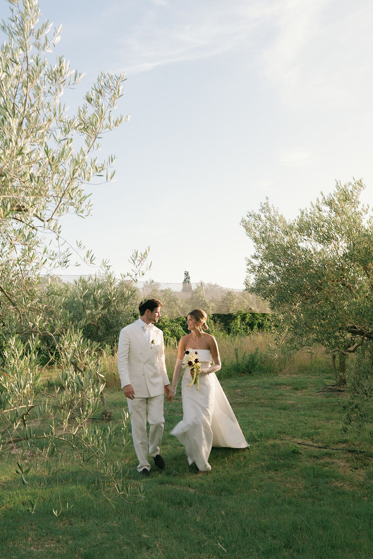 The bride and groom walking through long grass between olive trees
