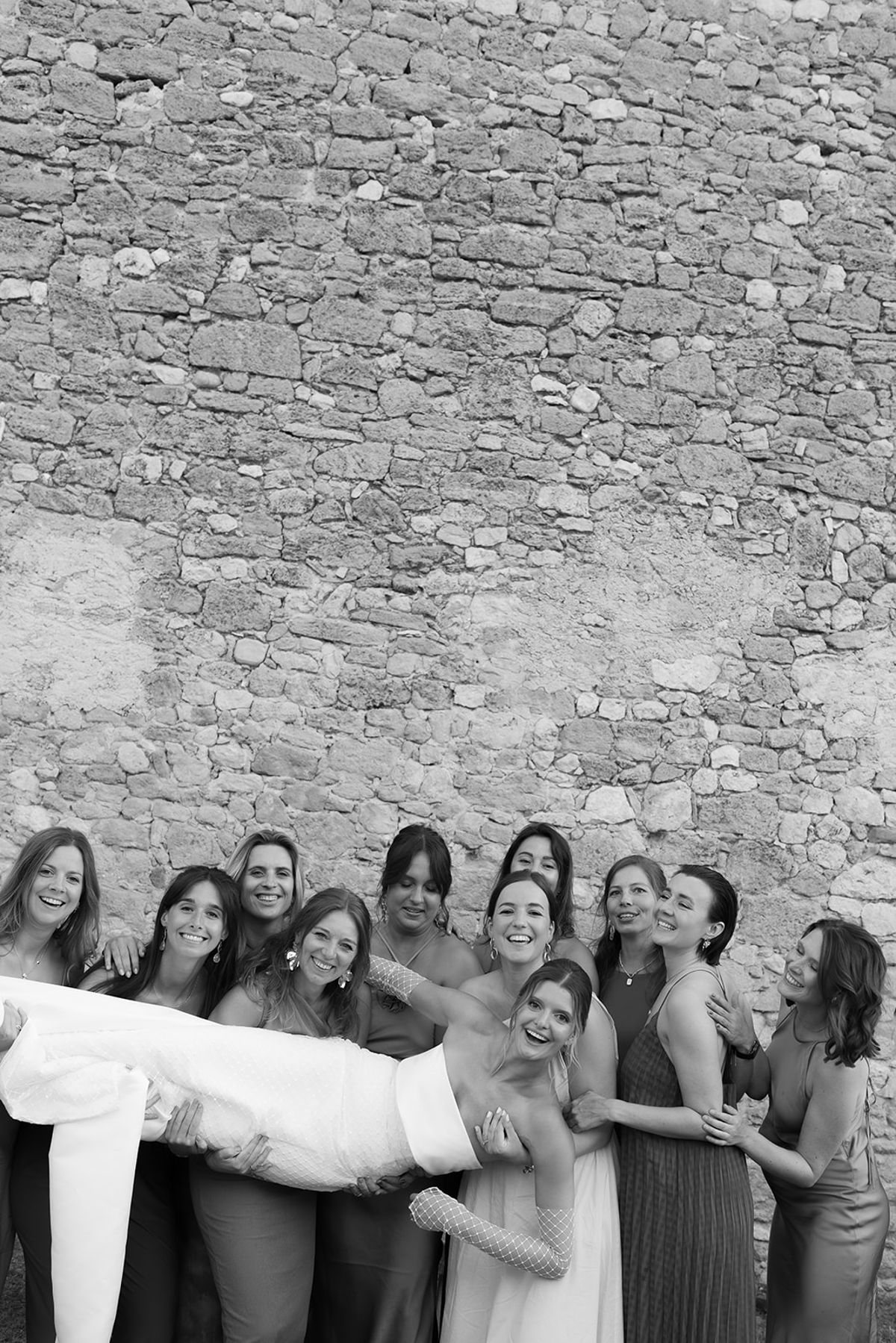 The bride's friends lift her sideways in front of a stone wall
