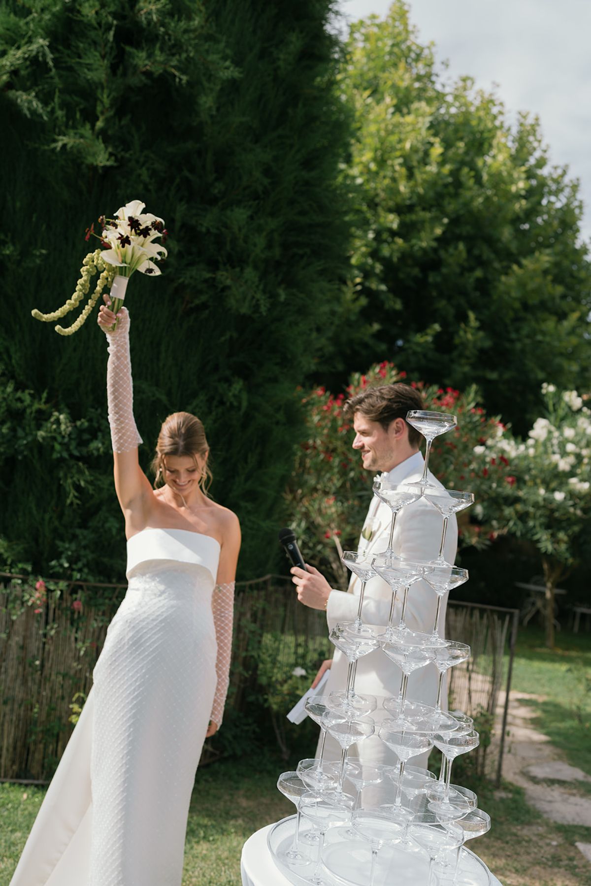 The bride raises her bouquet beside the champagne fountain as the groom looks on