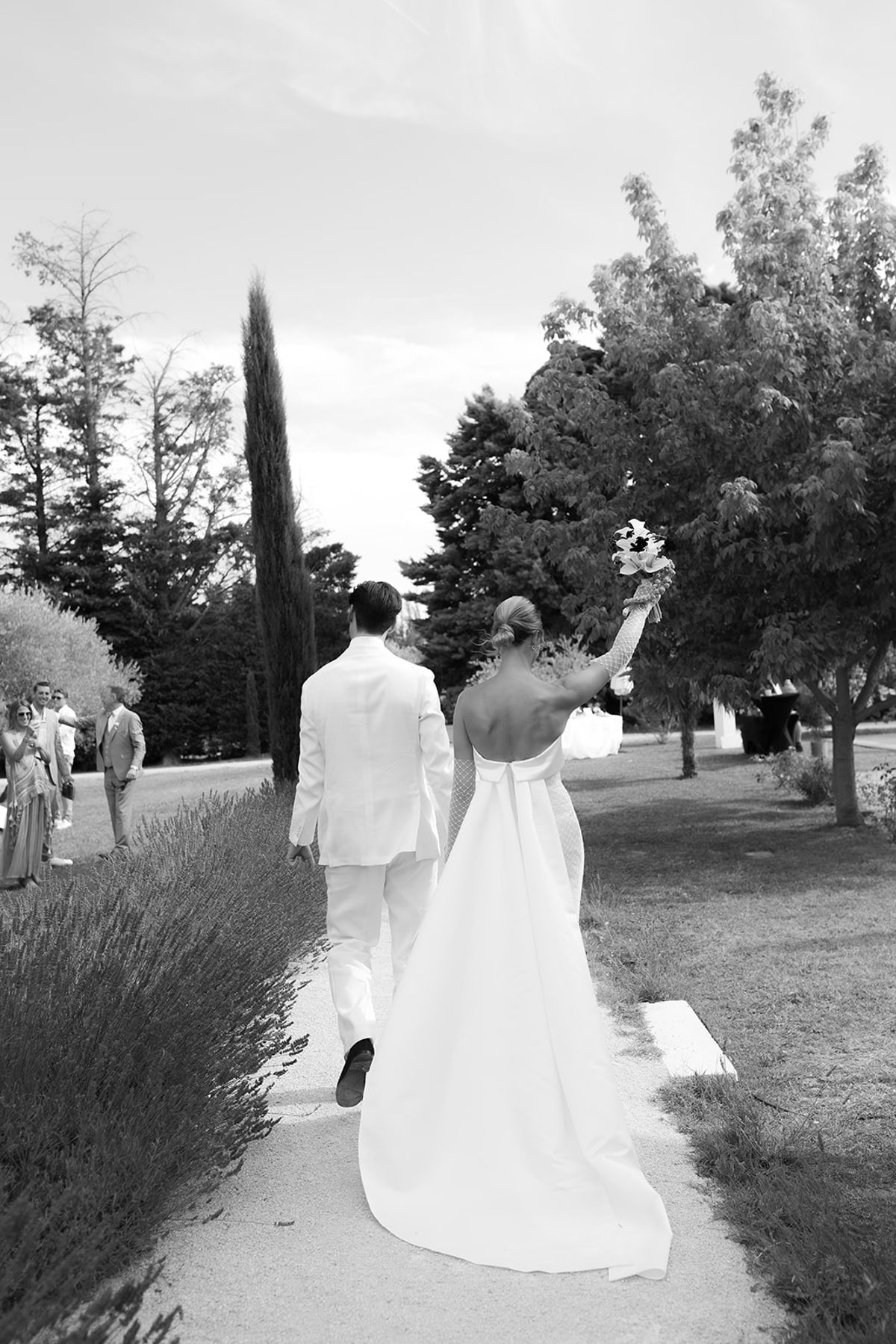 The bride and groom walk away along a path lined with lavender, her bouquet raised