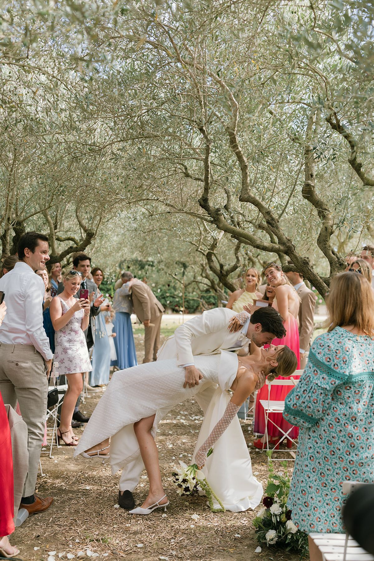 The groom dips the bride for a kiss as guests cheer in the olive grove