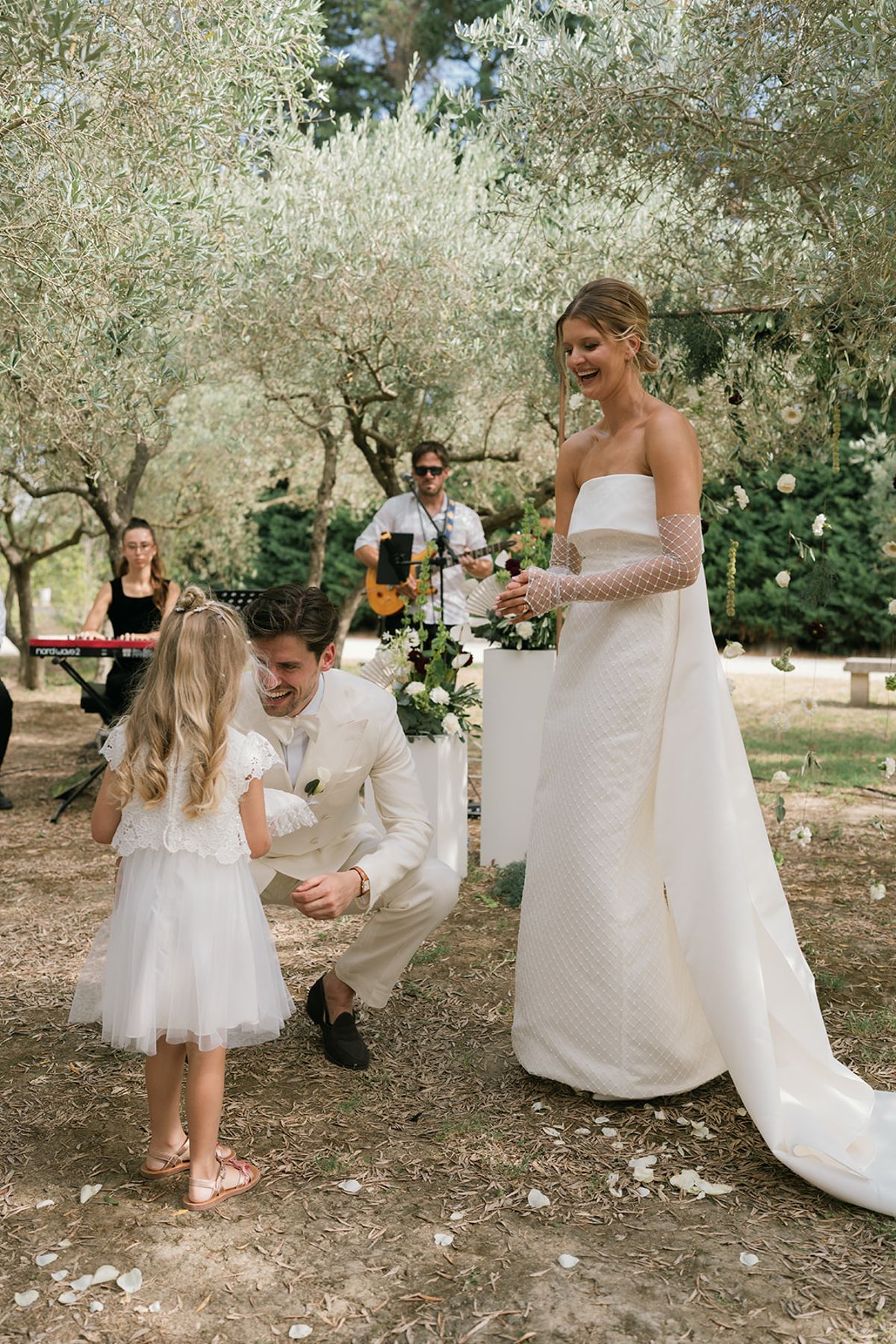 The groom kneels beside a flower girl as the bride smiles, with musicians playing behind them