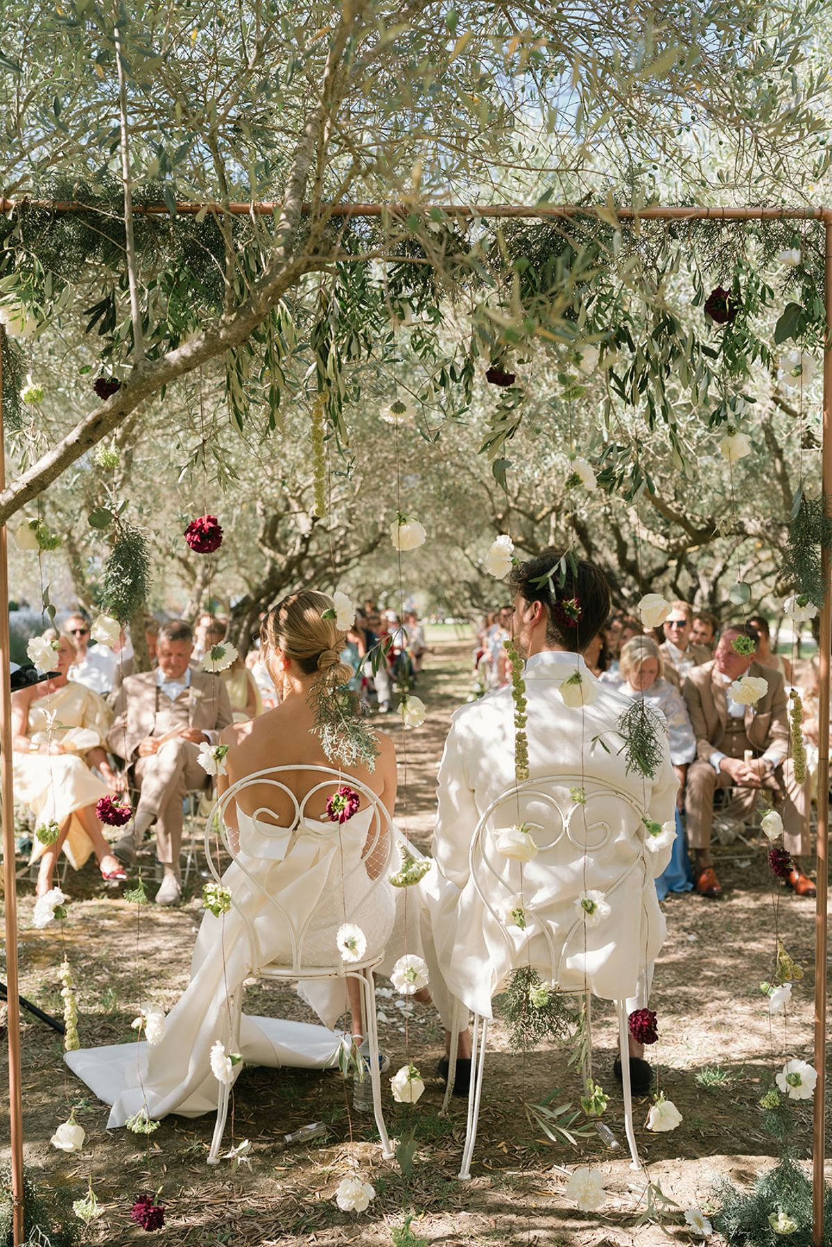 The ceremony seen from behind the guests, with dark red flowers hanging in the olive branches