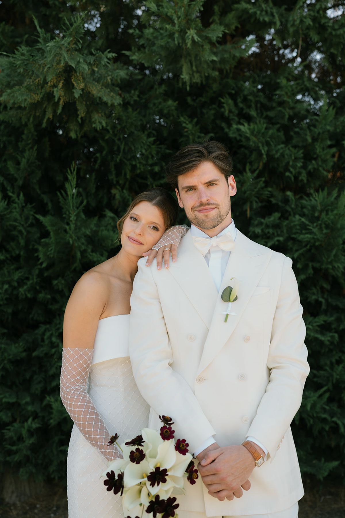 The bride resting her head on the groom's shoulder in front of a cypress hedge