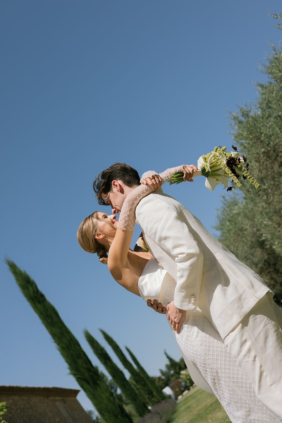 The groom lifts the bride's bouquet high as they kiss beneath cypress trees and a blue sky