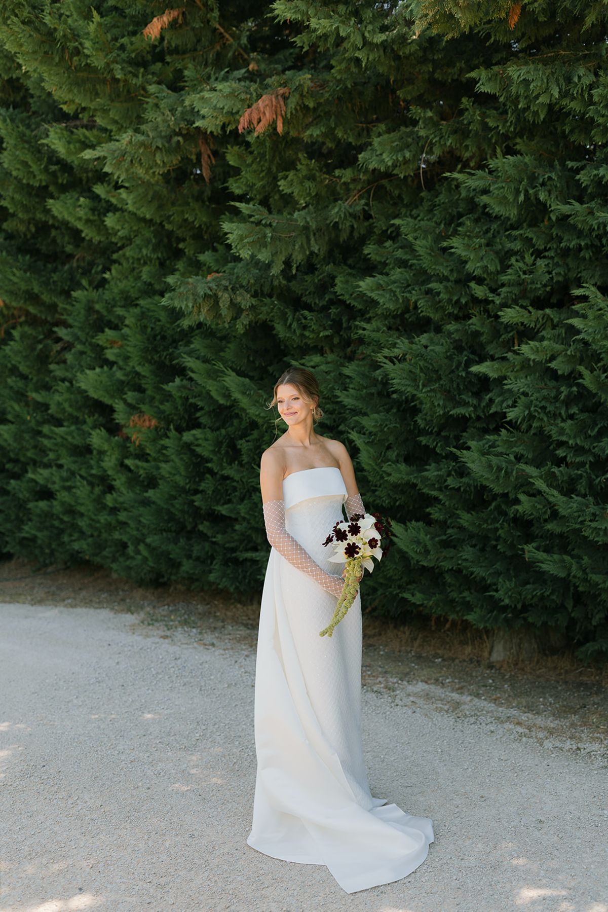 The bride standing alone on a gravel path in front of a tall green hedge