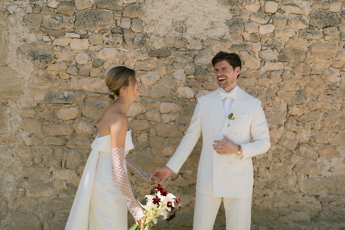 The bride and groom laughing and holding hands in front of an old stone wall at Le Petit Roulet