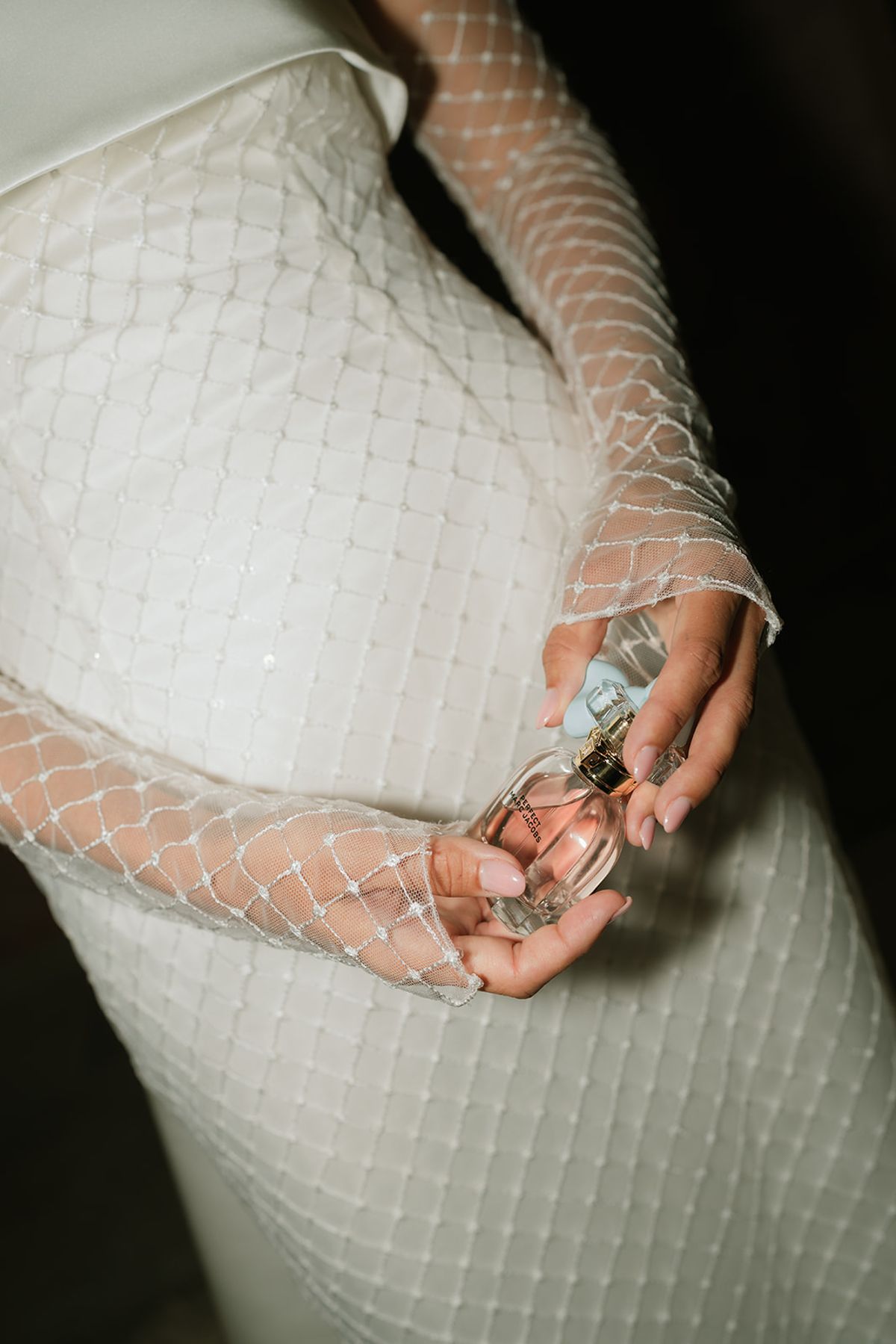 The bride's gloved hands holding a perfume bottle against the grid-pattern fabric of her dress