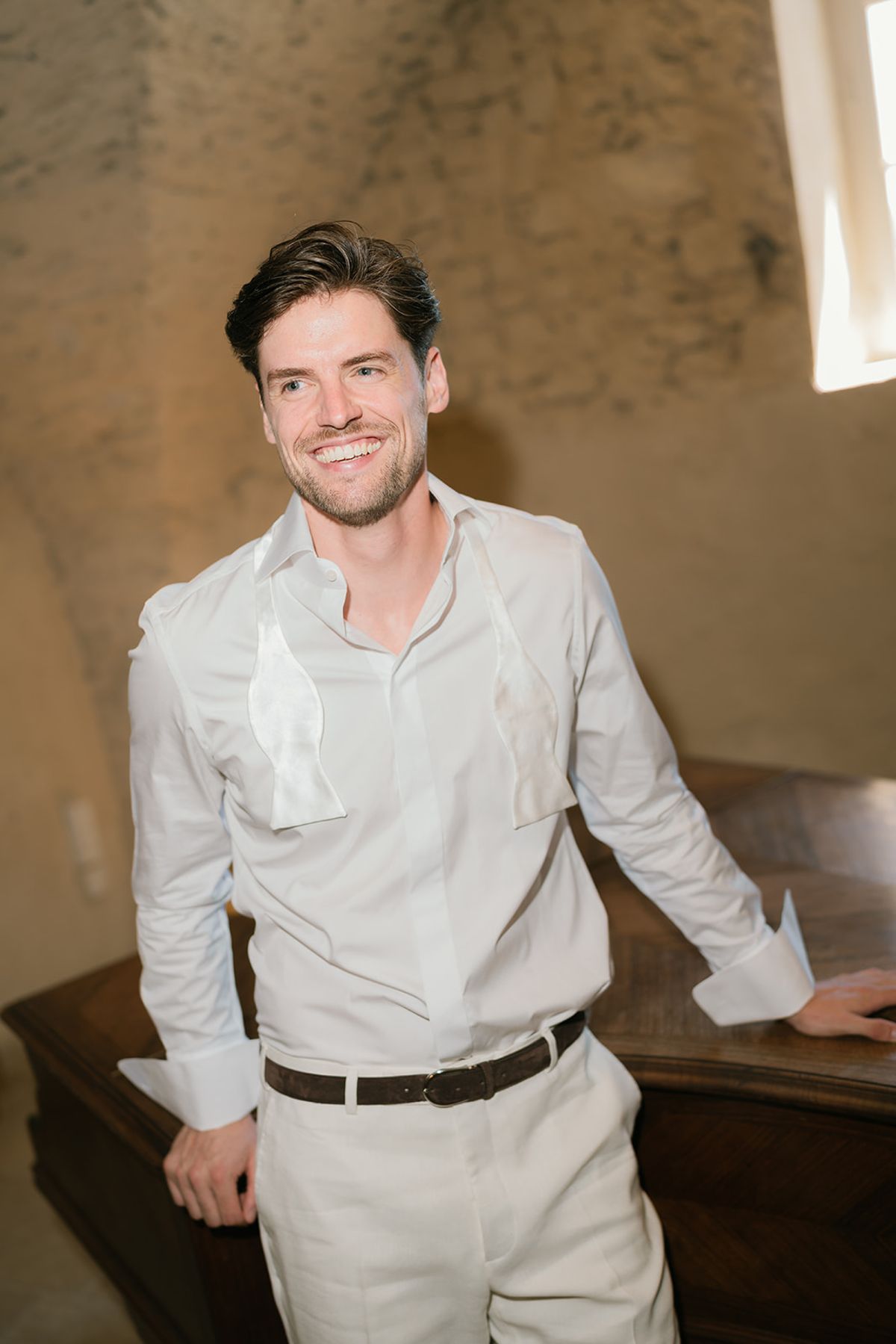 The groom in a white shirt and cream trousers in a vaulted stone room