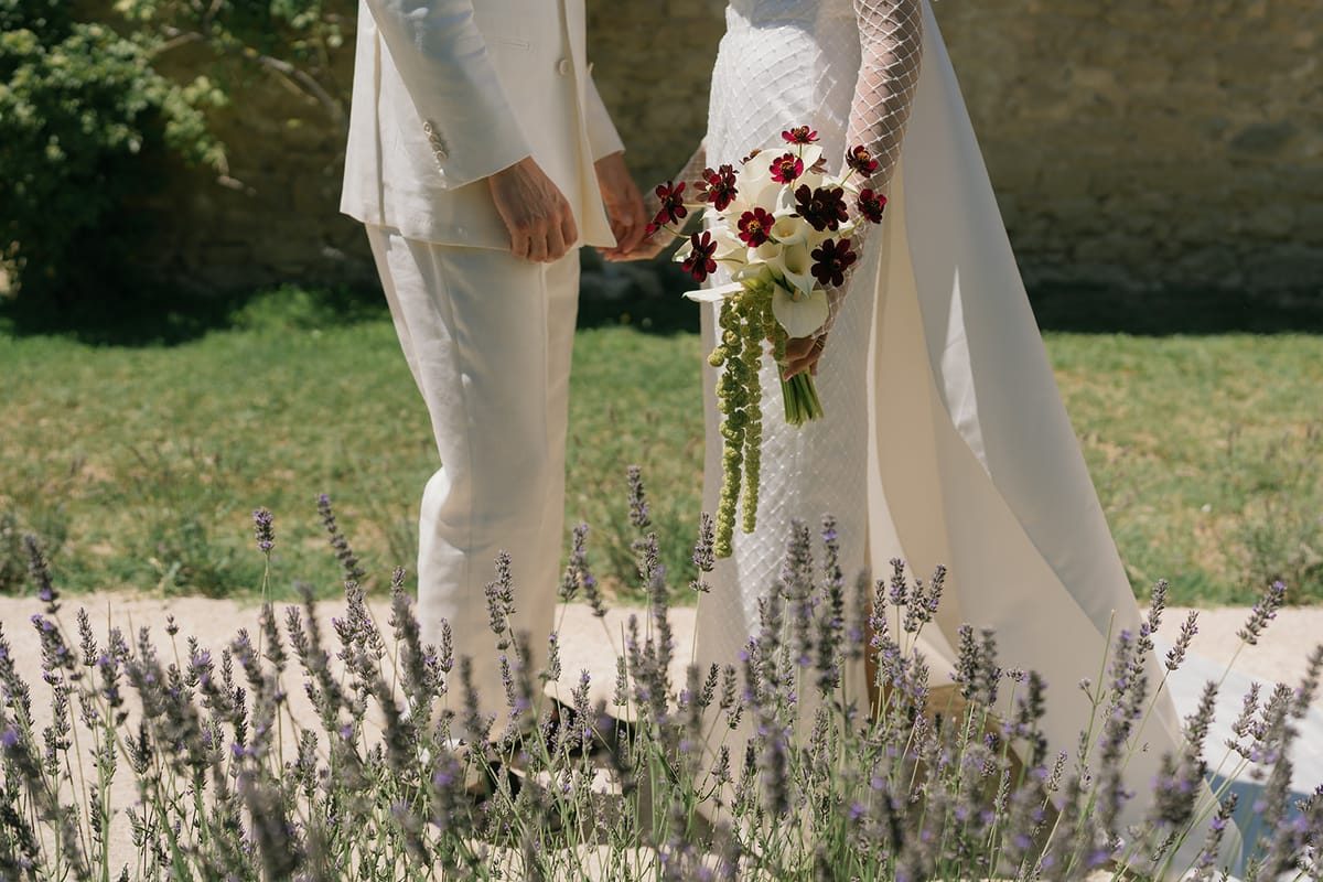 The bride and groom holding hands among the lavender, her bouquet of calla lilies and dark red cosmos at her side