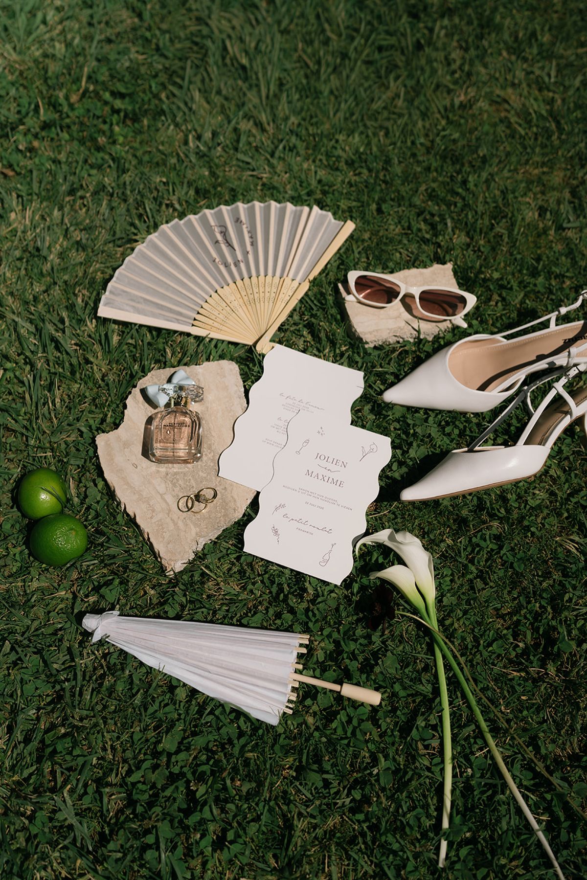 A flat lay on the grass of invitations, a calla lily, a folding fan, sunglasses, perfume, shoes and a white parasol