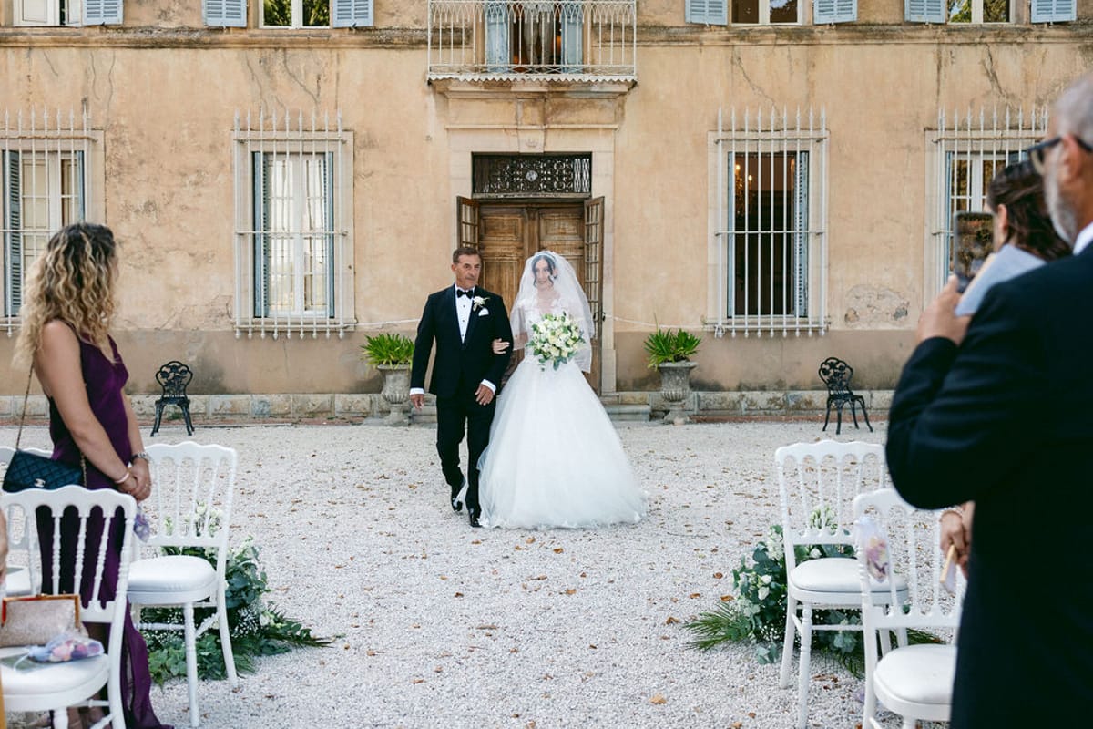The bride walking toward the groom on the gravel courtyard as guests look on