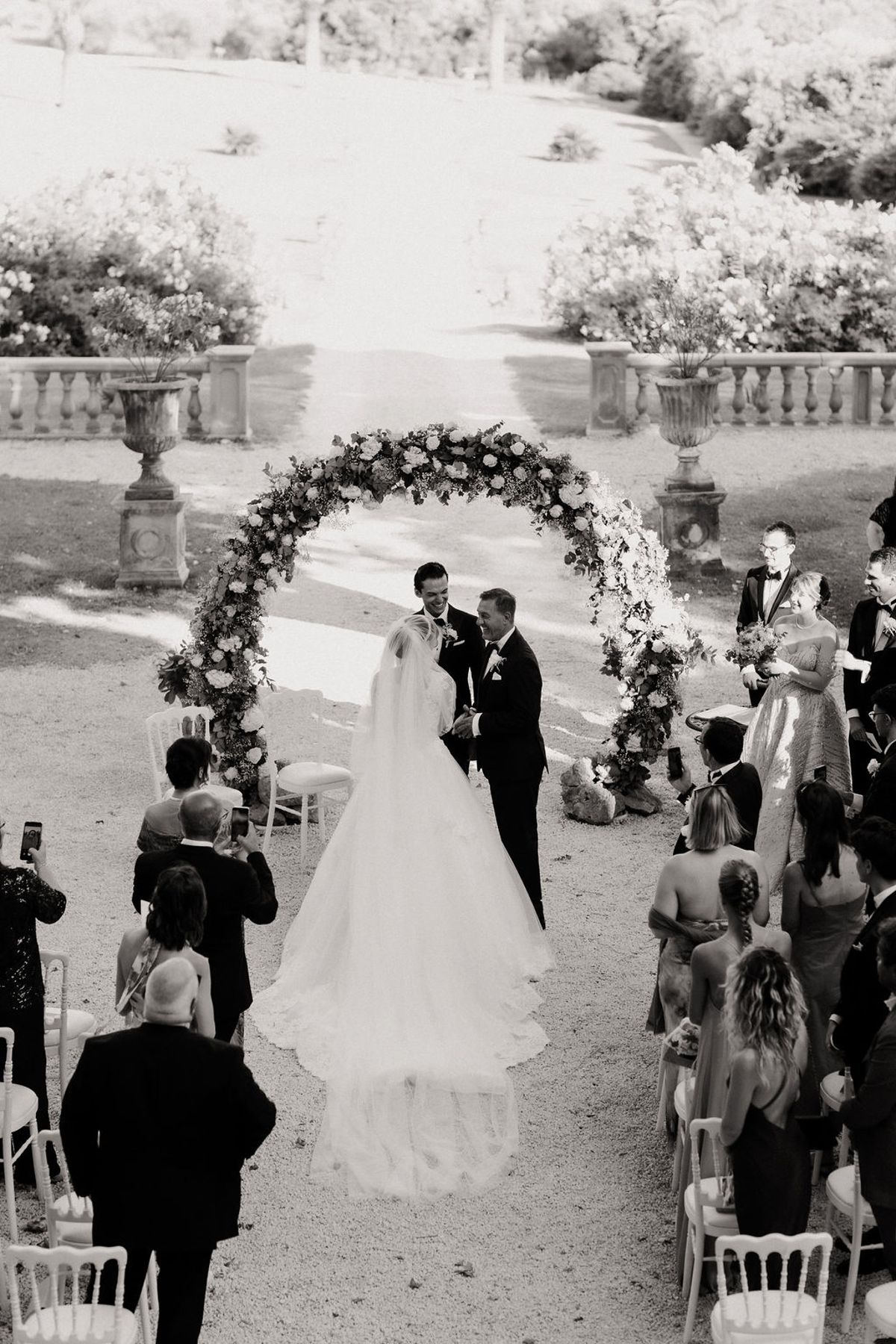 Black and white photograph of the bride walking the aisle toward the floral arch