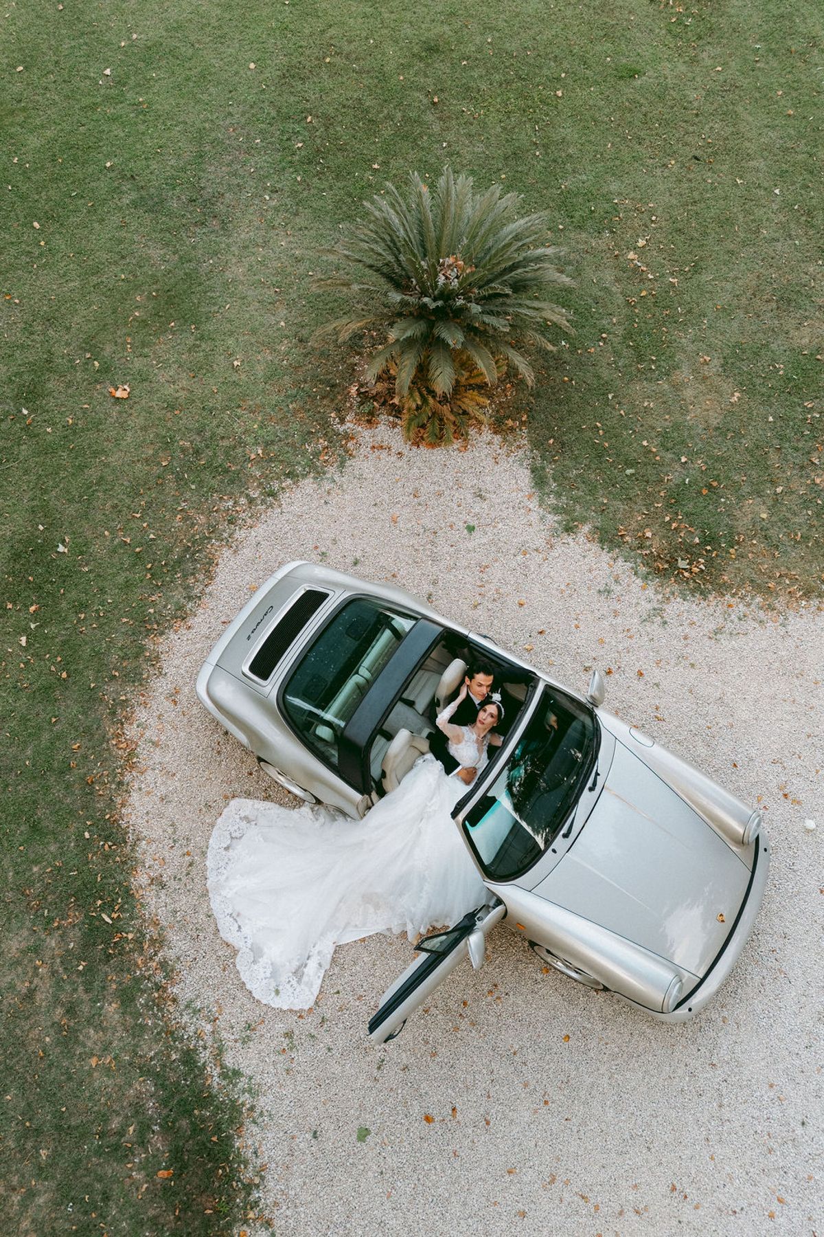 Aerial photograph of Ilenia and Dalil beside a white convertible on the gravel drive