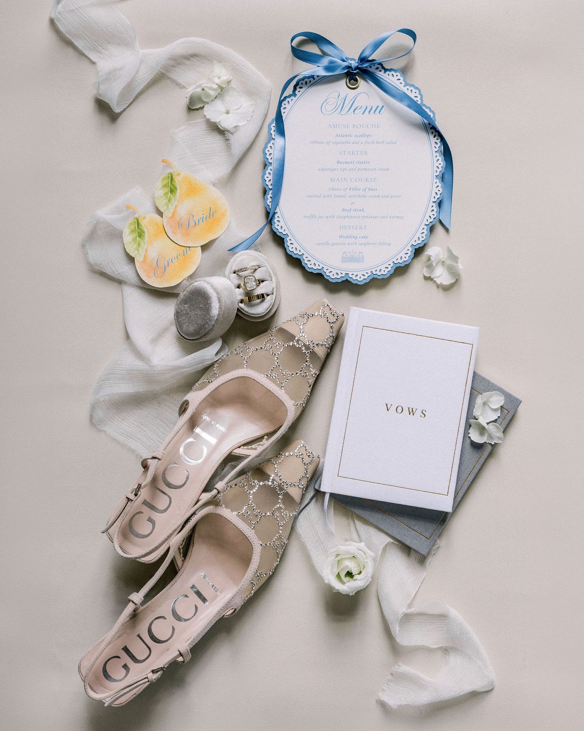 Flat lay of a blue scalloped menu tied with ribbon, a vows booklet, jewelled slingback heels and the rings