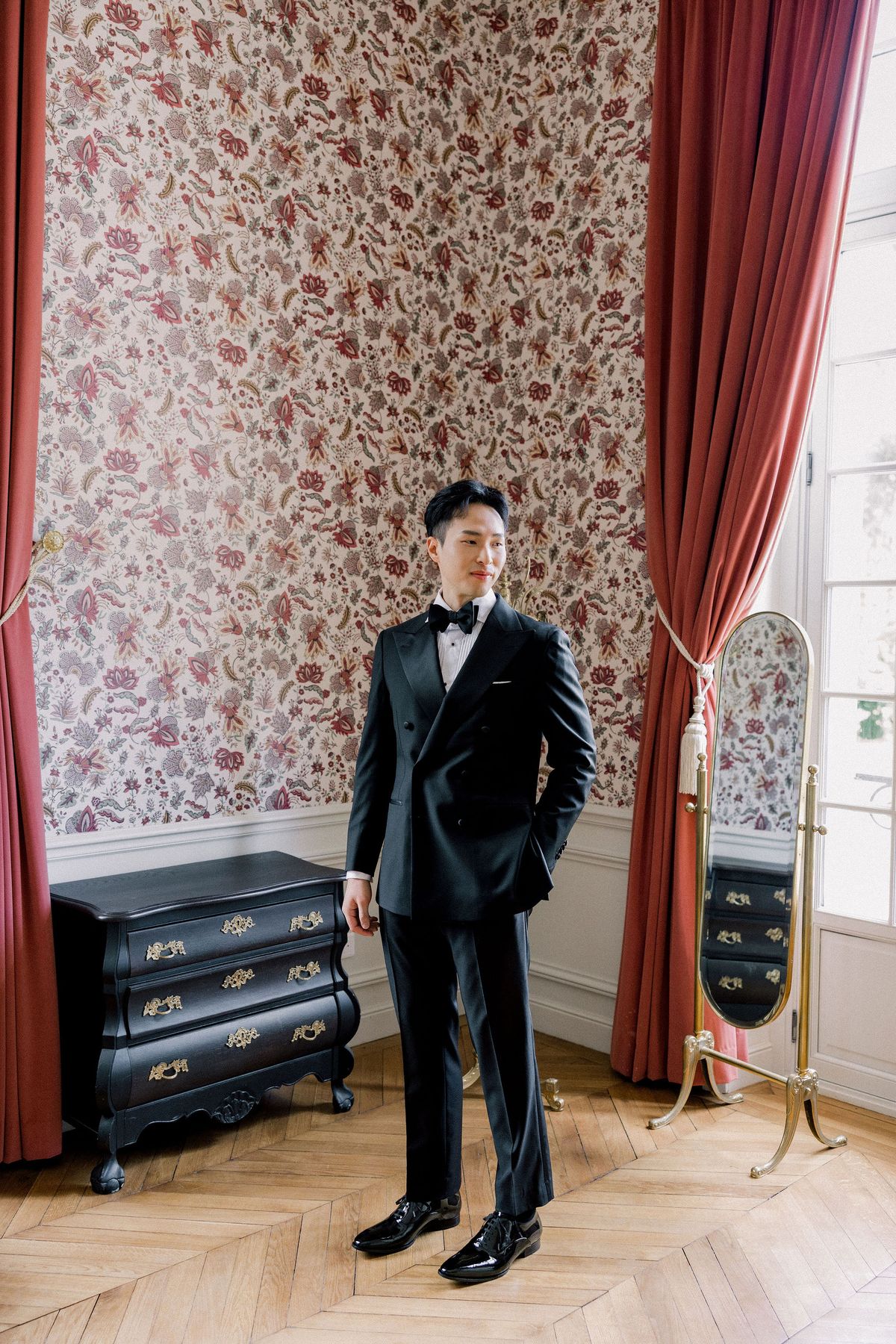 The groom in a black tuxedo stands in a room hung with red floral wallpaper