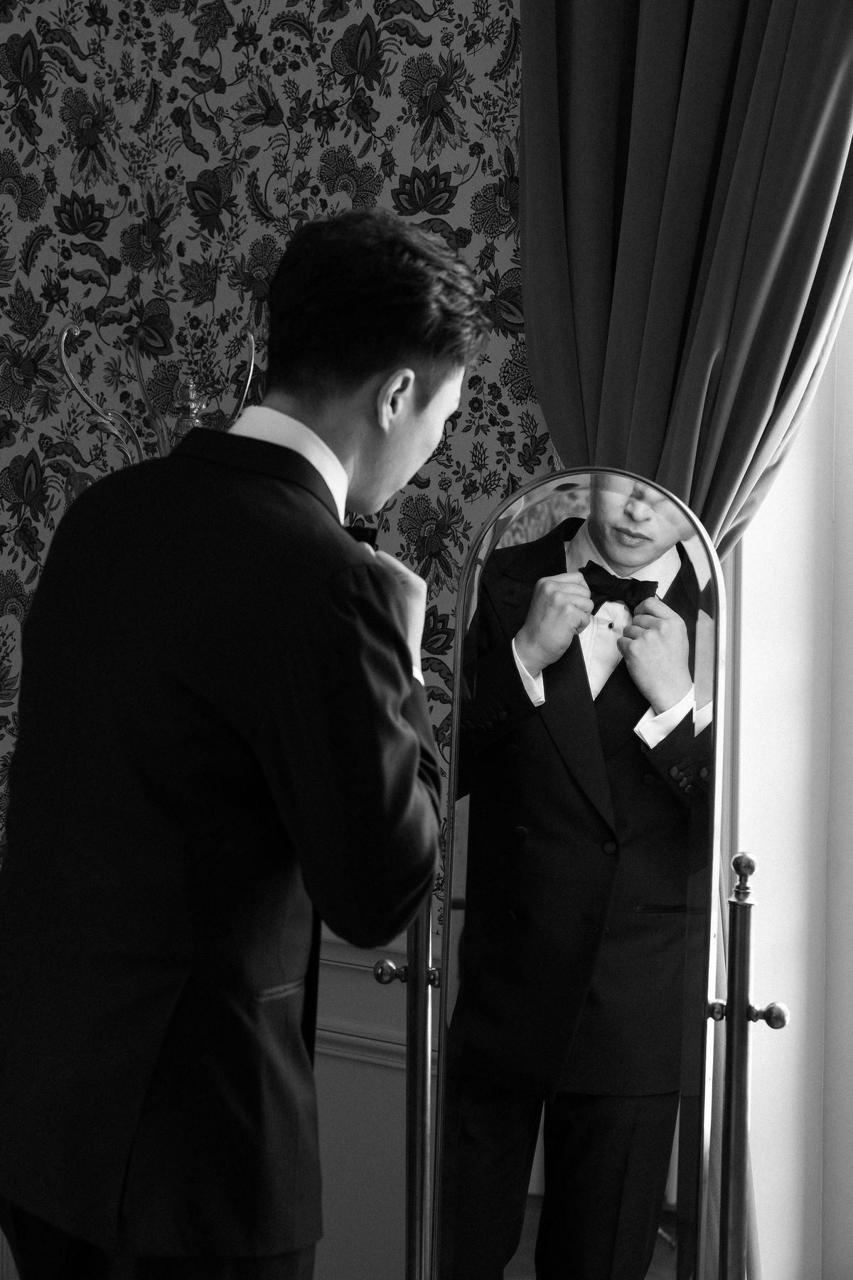 Black and white image of the groom adjusting his bow tie in a standing mirror