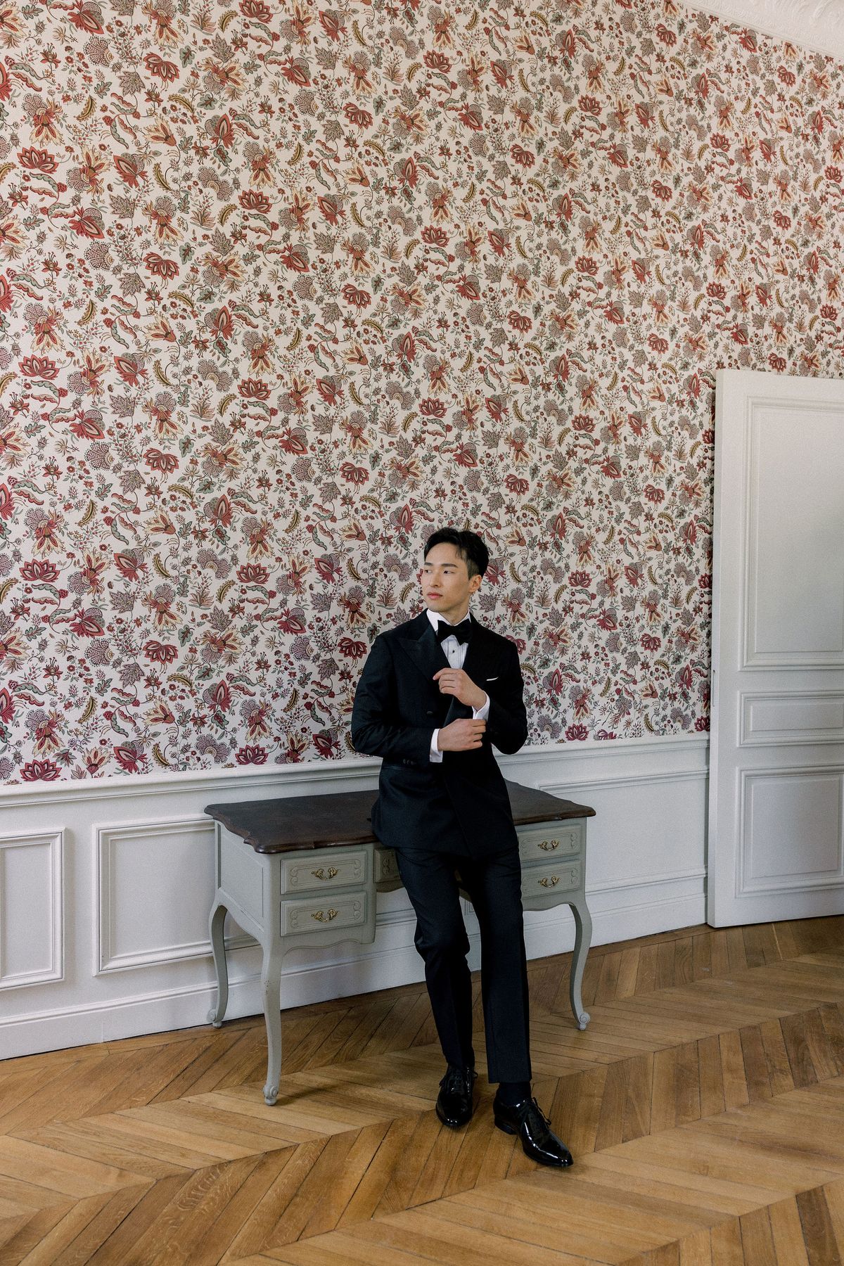 The groom leans against a grey console table in a room with red floral wallpaper