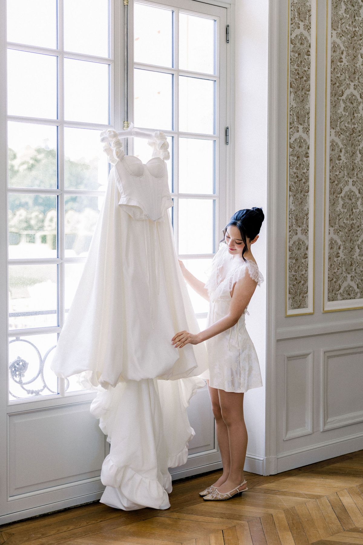 The bride adjusts her wedding gown hanging in a tall window of a panelled salon
