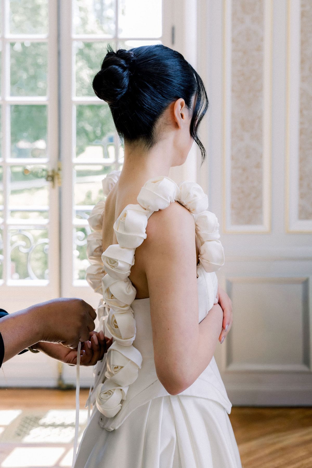 The bride is laced into her gown, sculpted fabric roses running down the back of the straps