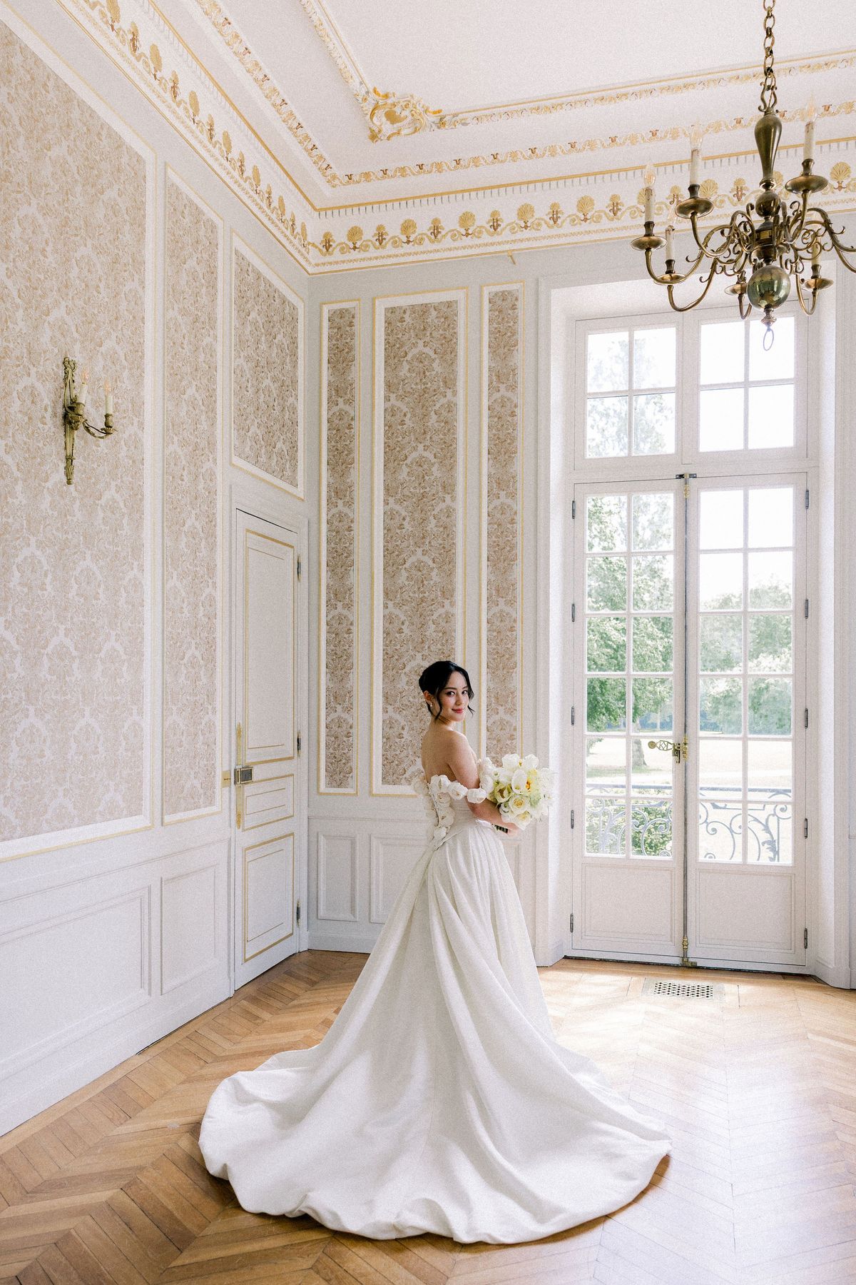 The bride stands in a white and gold salon beneath a brass chandelier, her train spread on the parquet